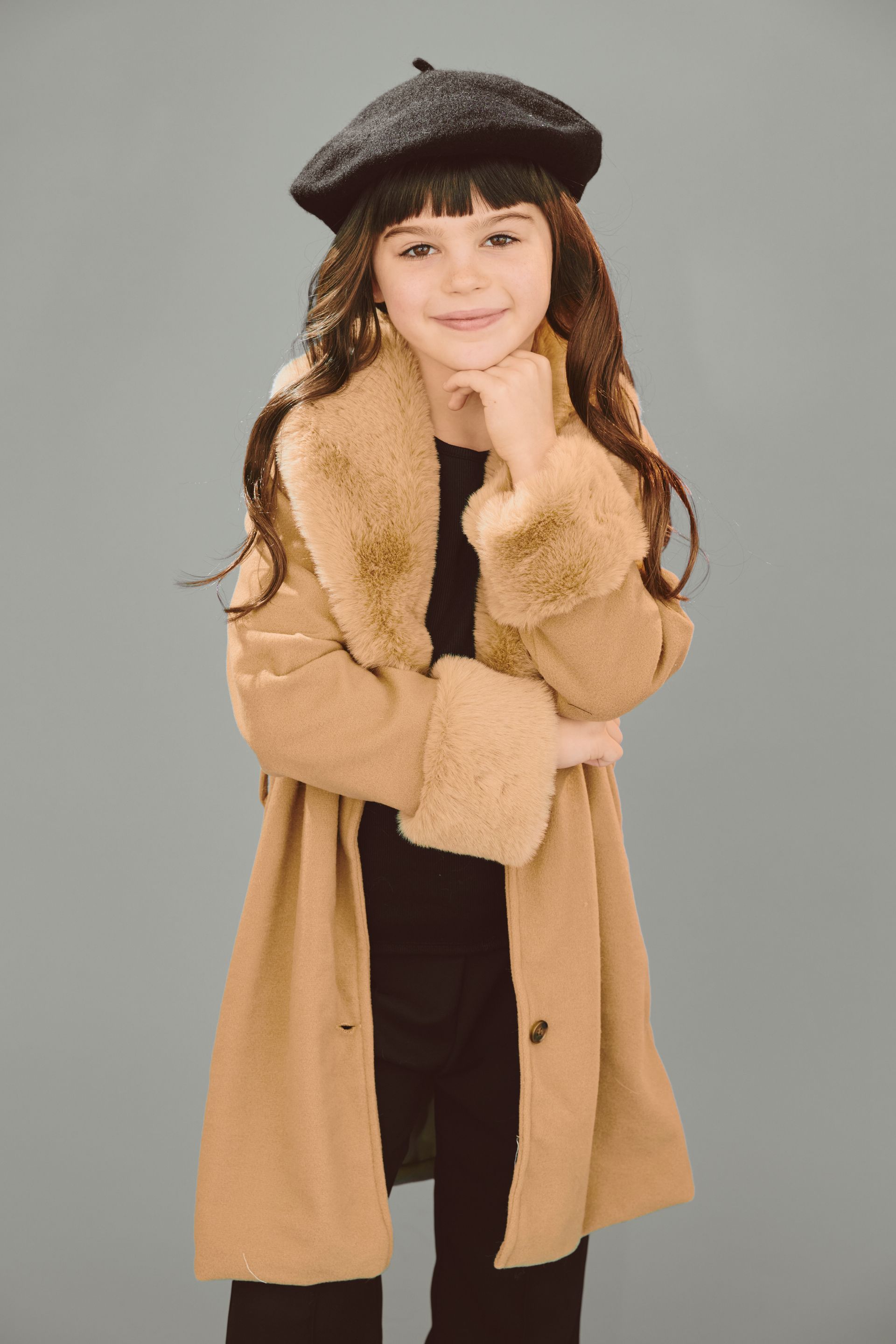 Girl wearing a tan coat and black beret, with hand on chin, smiling. Against a gray background.