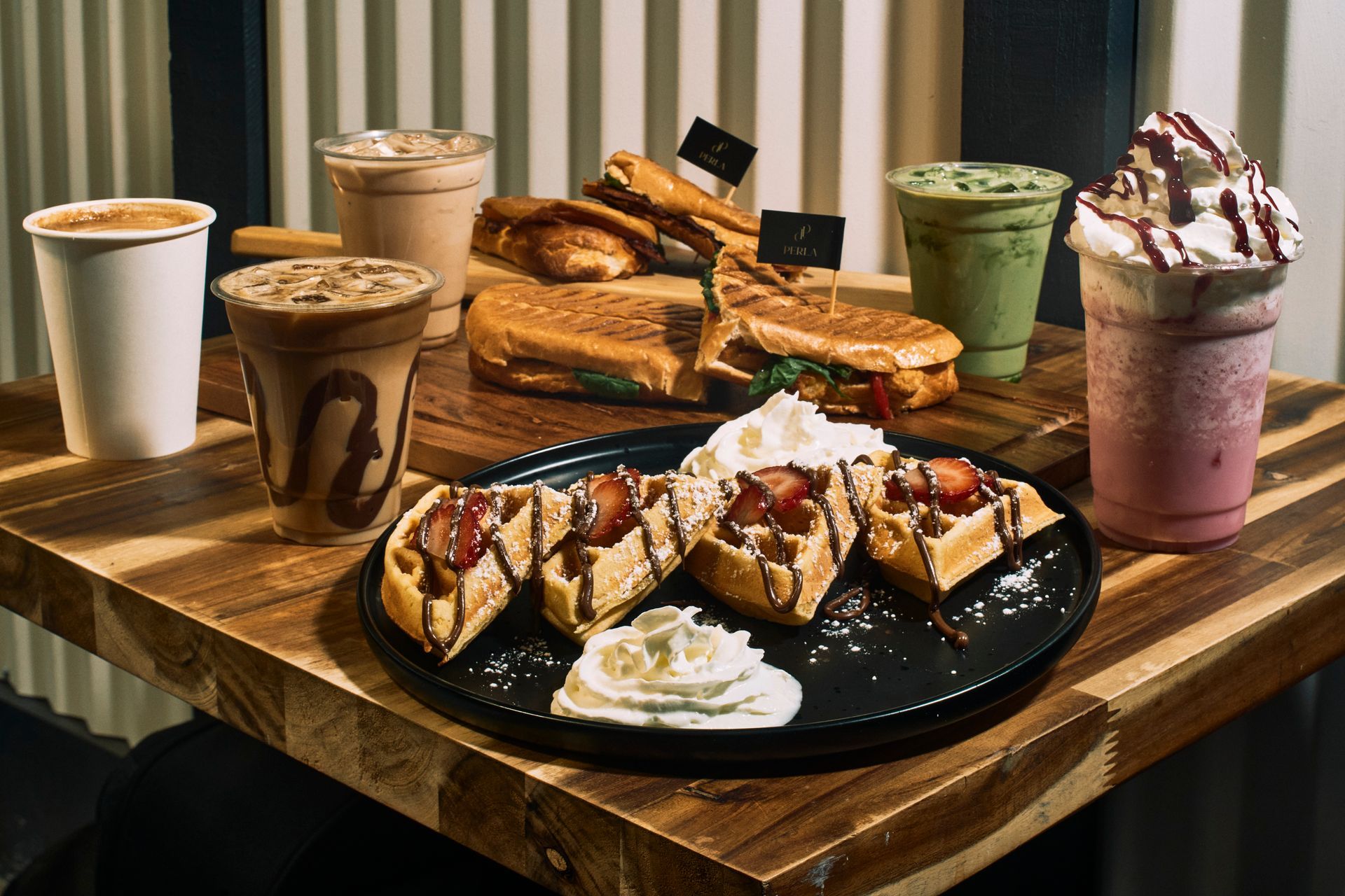 Waffles with strawberries and whipped cream, sandwiches, and assorted drinks on a wooden table.
