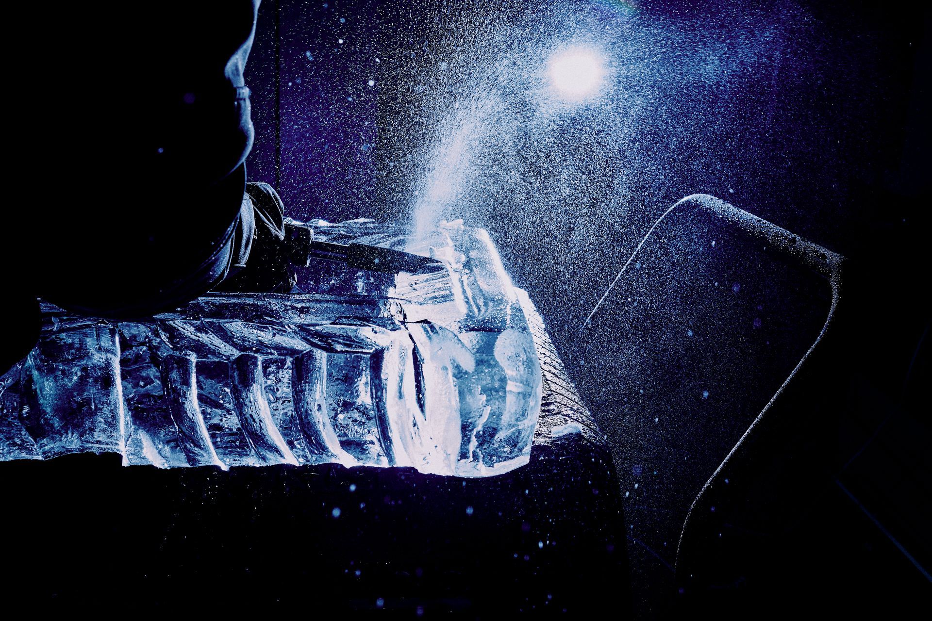 Person breaking ice with a tool, lit by a bright light. Blue tones, dark setting.