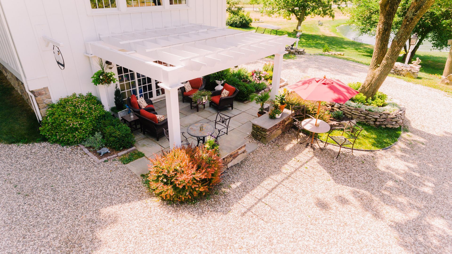White pergola over patio with seating, surrounded by gravel, plants, and small umbrella.