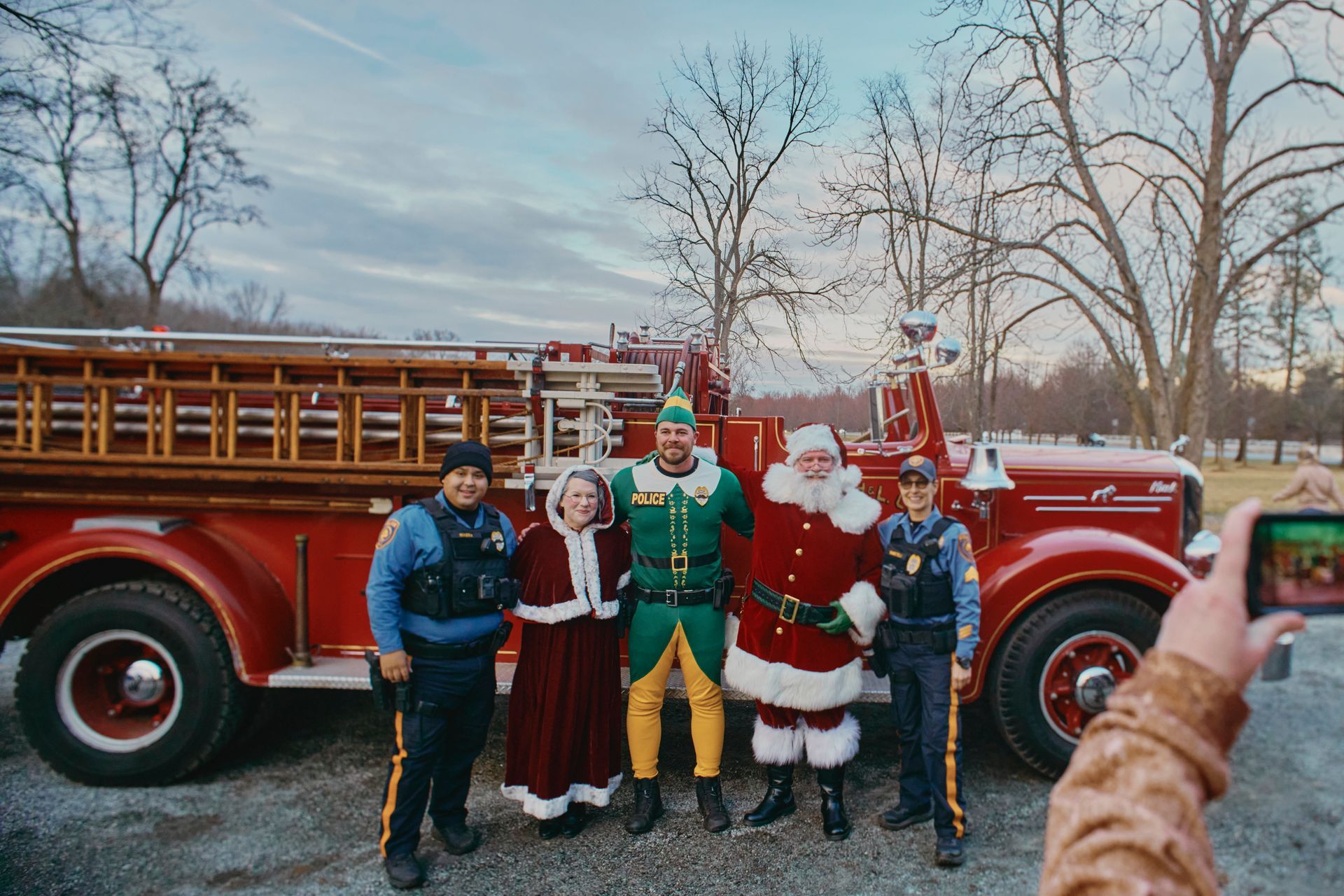 People in costumes pose with a vintage fire truck. Santa, an elf, a woman, and two officers smile.