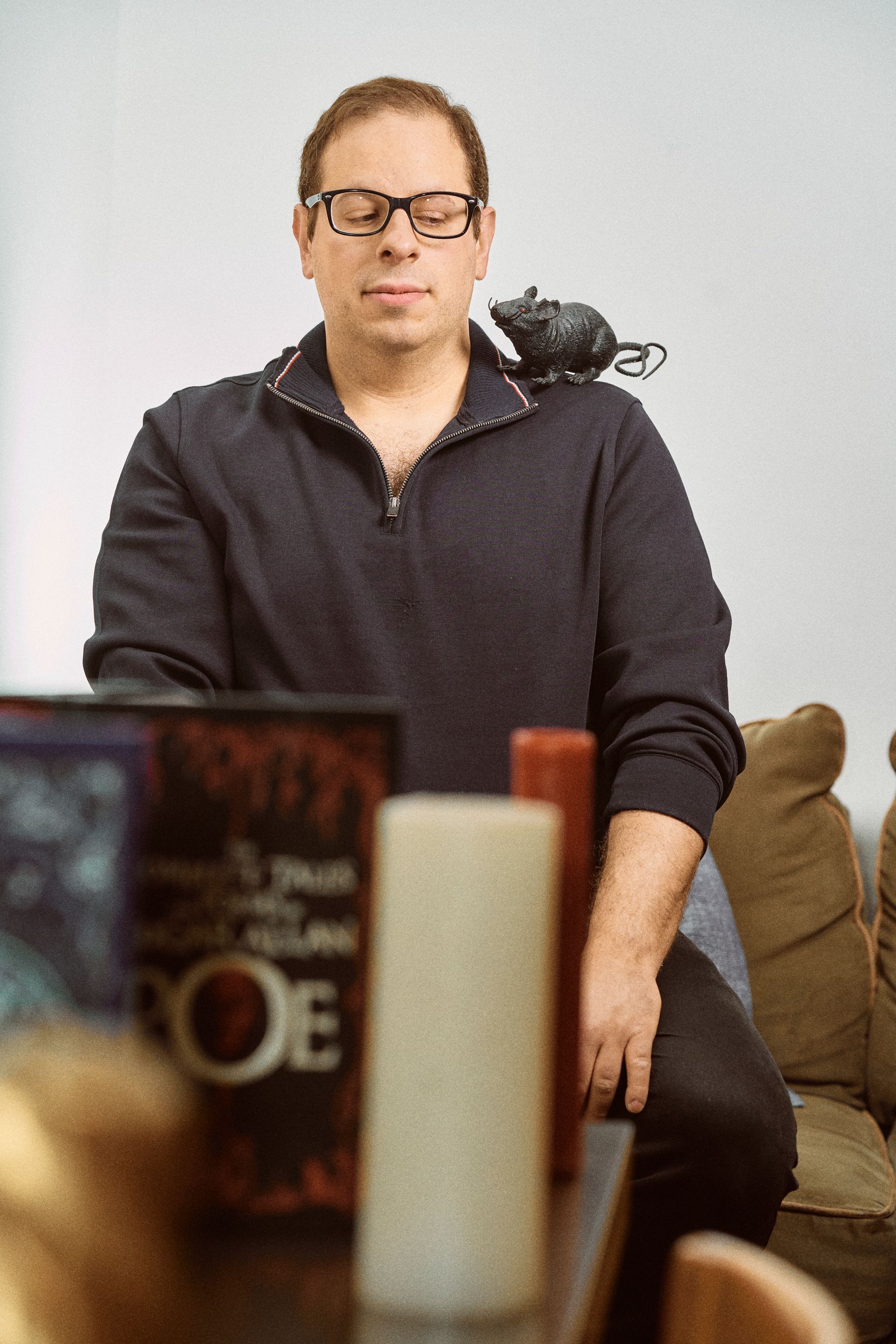 Man with glasses and a small rat on his shoulder, sitting indoors near a table with books and candles.
