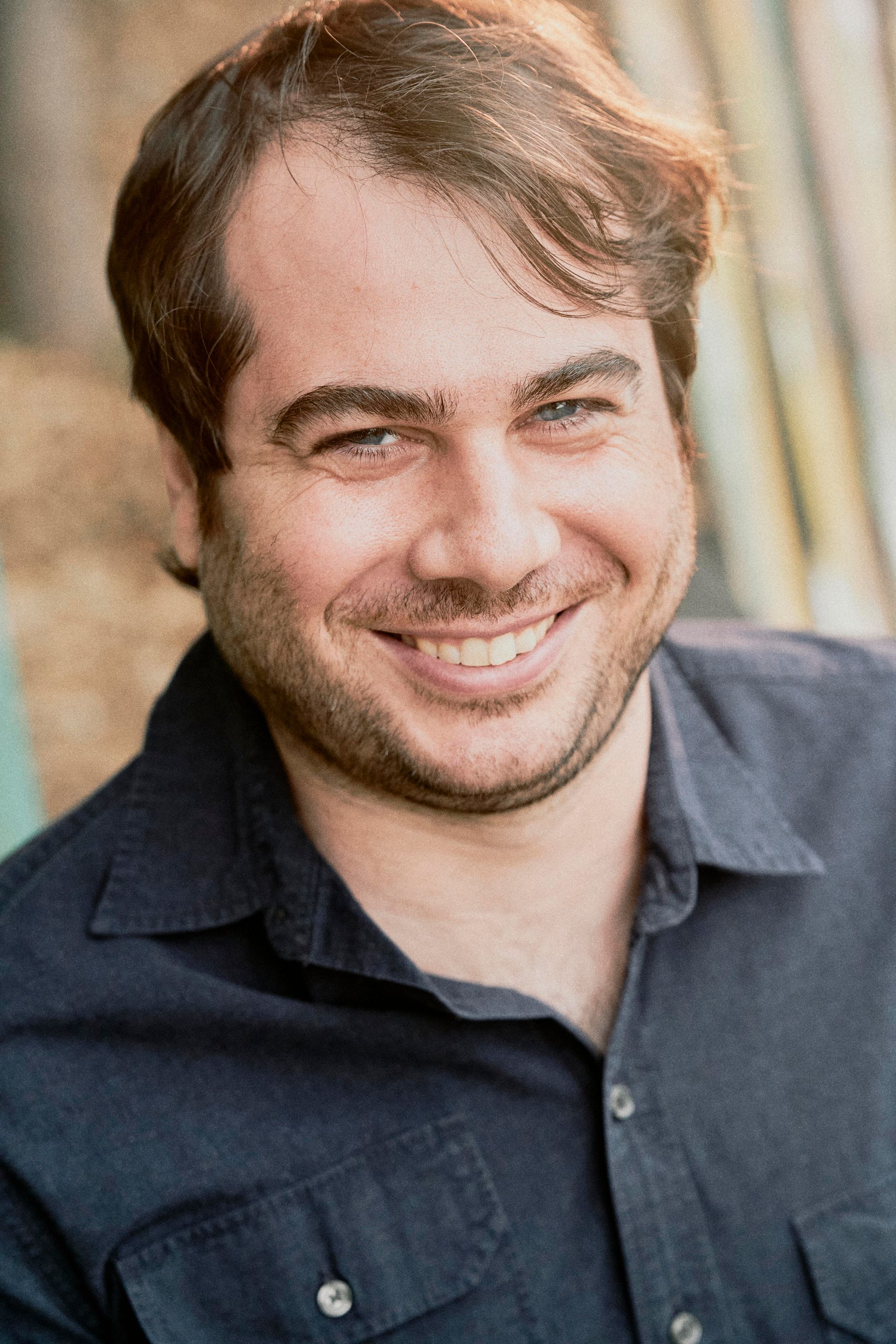 A man with a beard is smiling and wearing a blue shirt.
