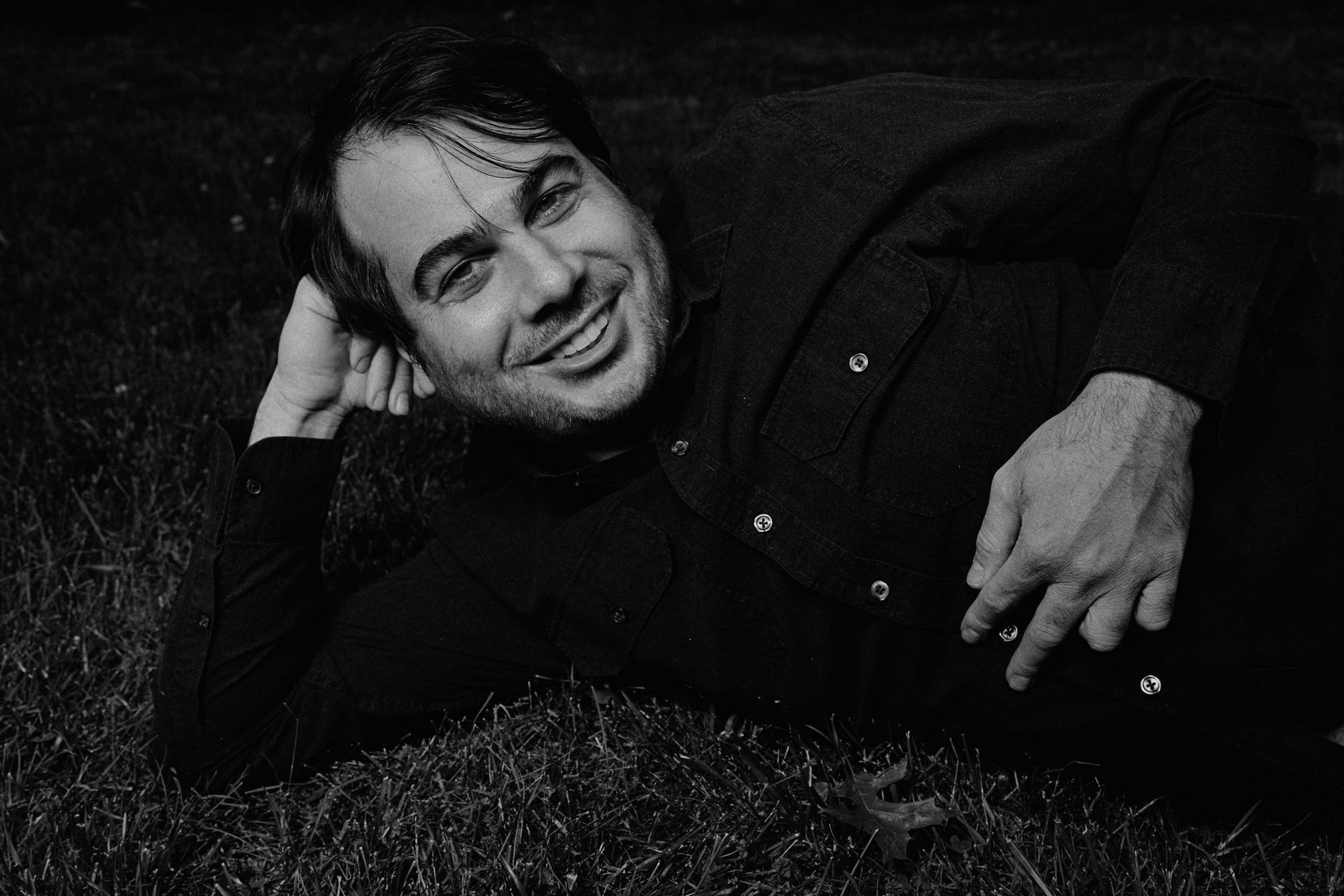 A man in a black shirt is laying on the grass and smiling.