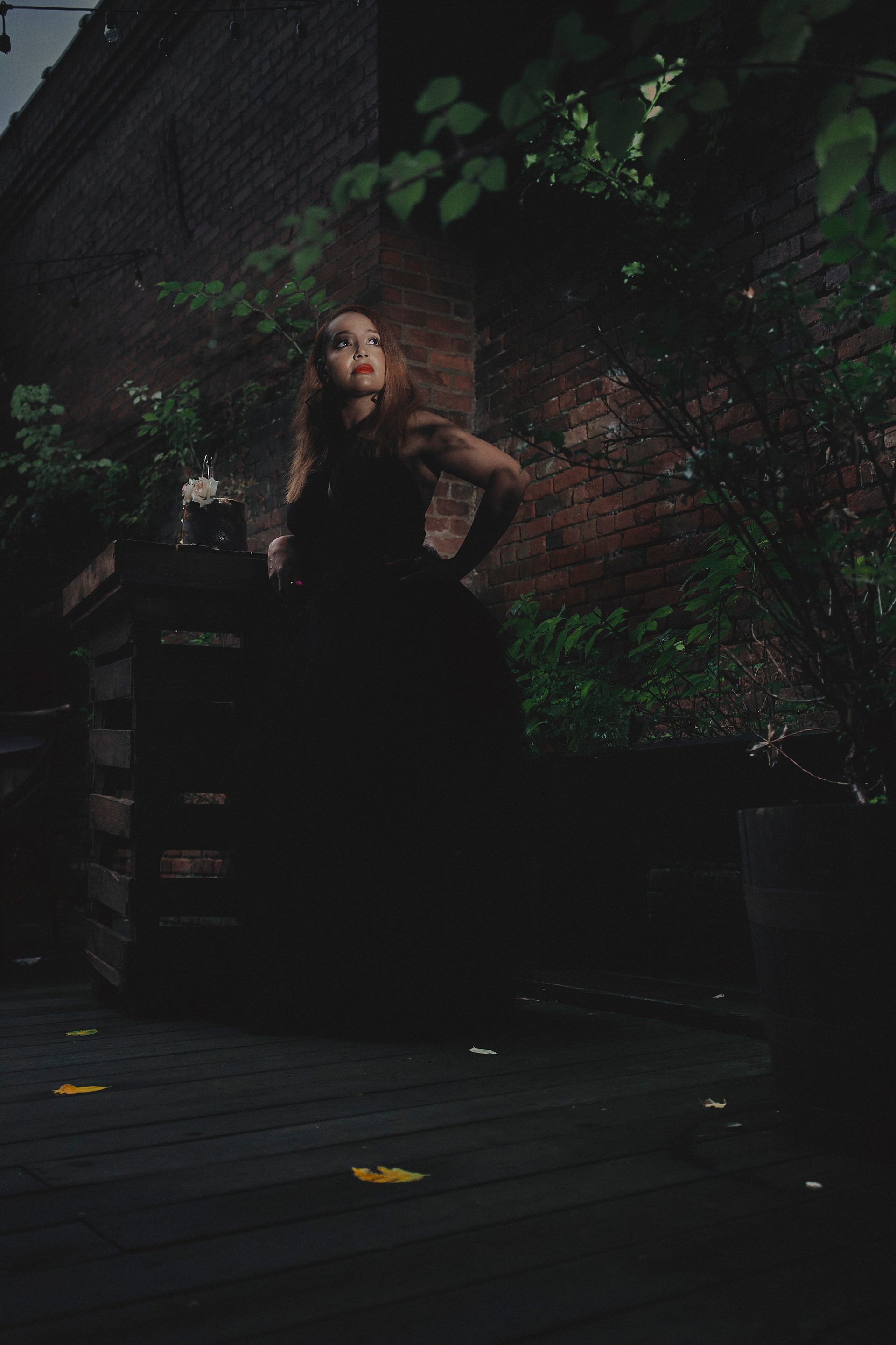 A woman in a black dress is standing in front of a brick wall.