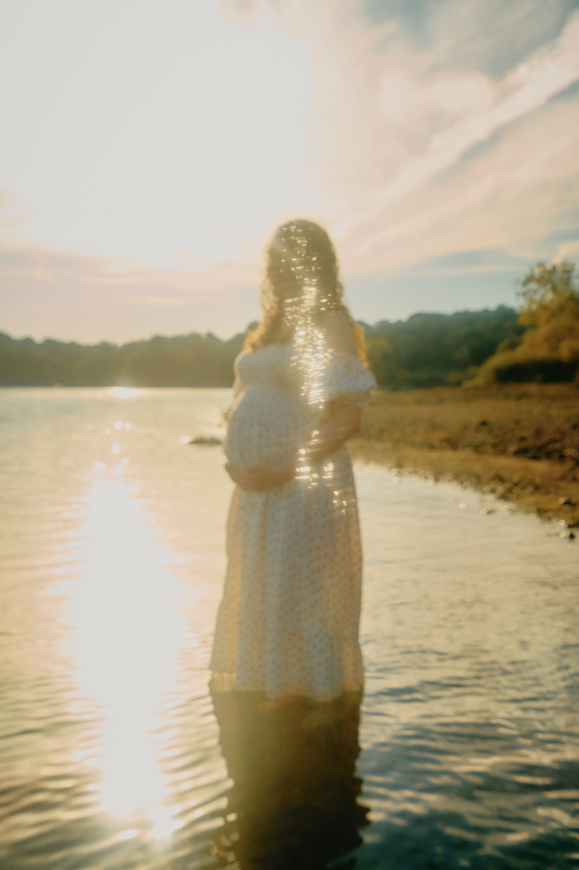 Pregnant person stands in water, sunlight shining. Wearing a white dress, hands on belly.