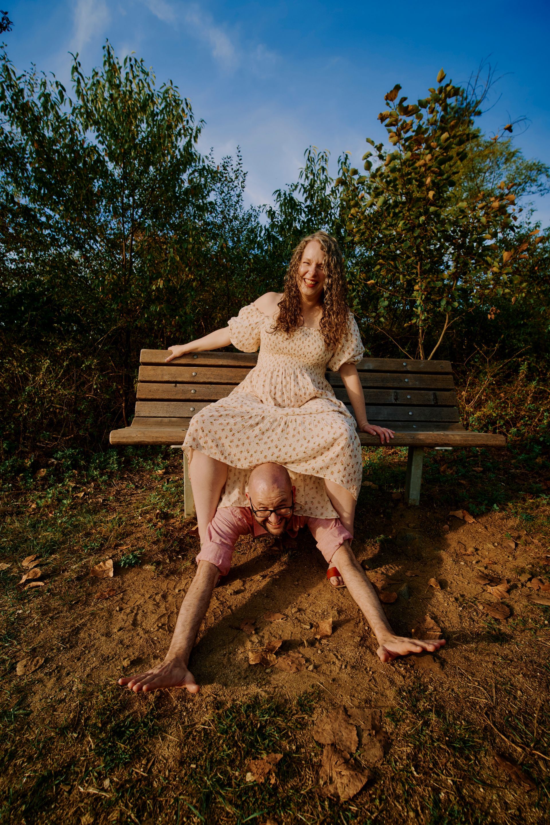 Woman in dress sitting on bench, man below with legs spread, outdoors.