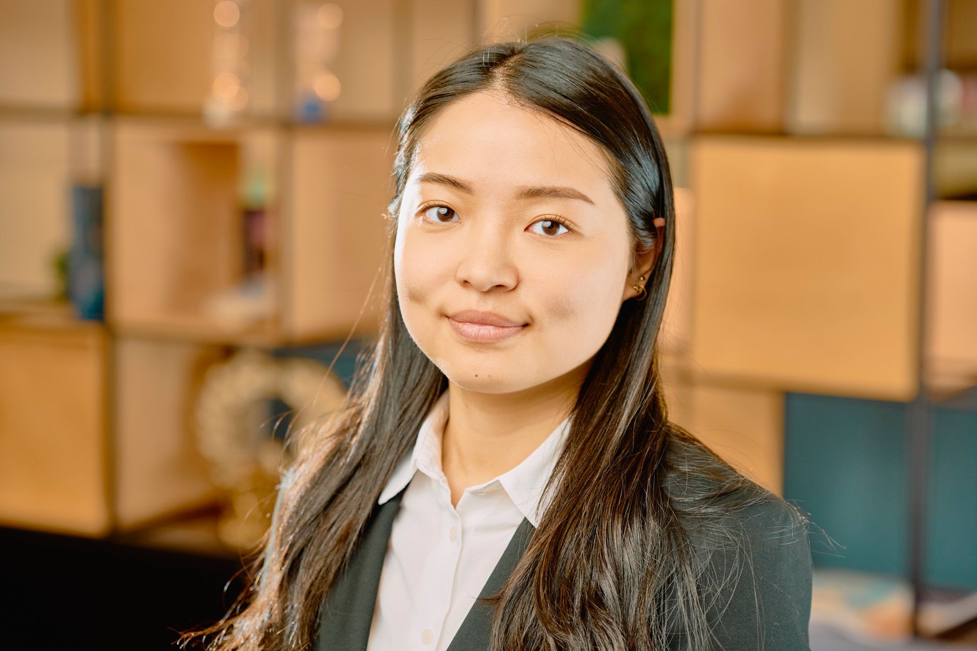 Woman in a black blazer and white shirt smiling in a warm office setting with shelves behind her