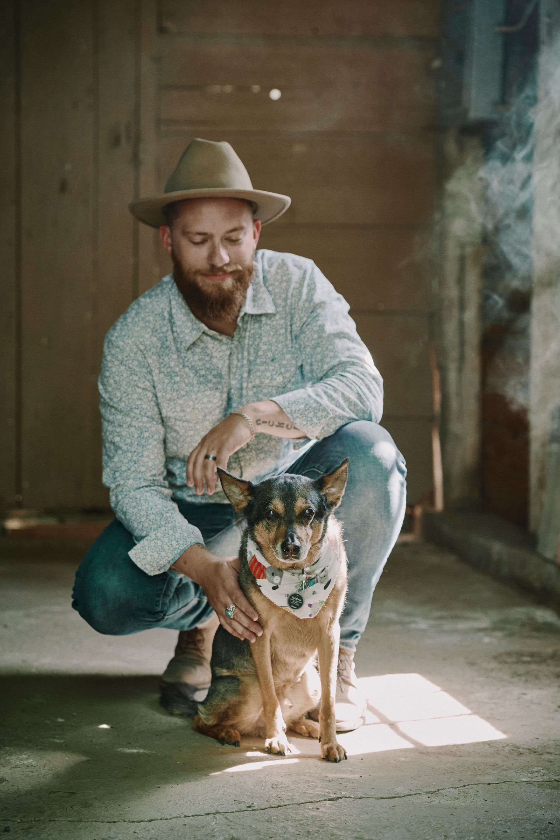 A man in a hat is kneeling down next to a small dog.