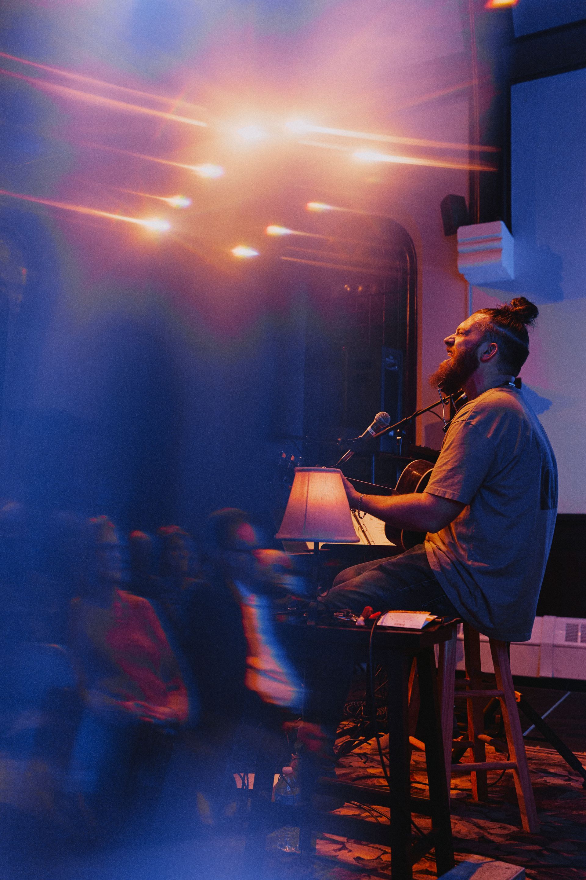 A musician with a top knot performs on a stool, singing into a mic. Stage lit with blue and orange.