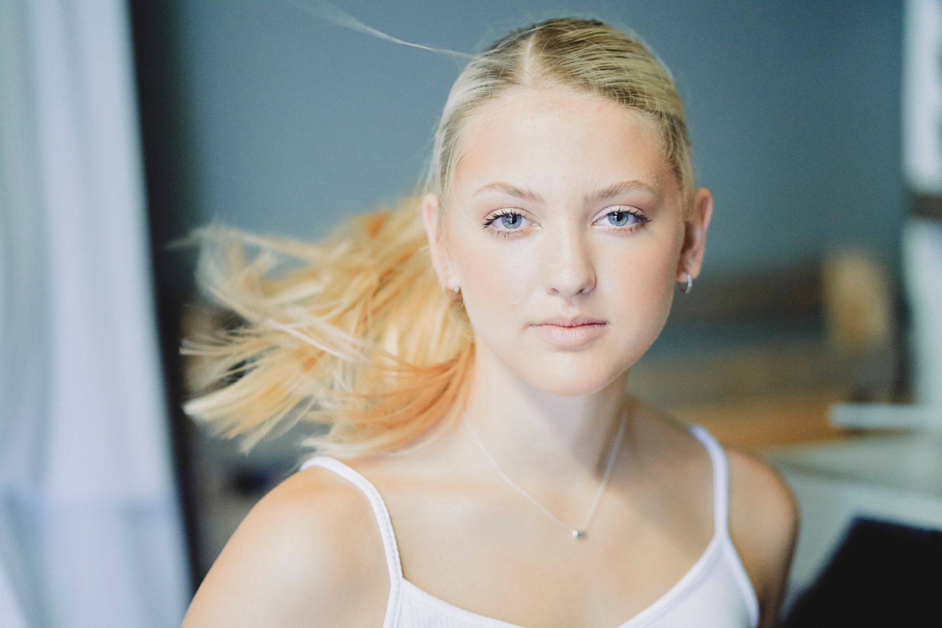A young woman in a white tank top is looking at the camera with her hair blowing in the wind.