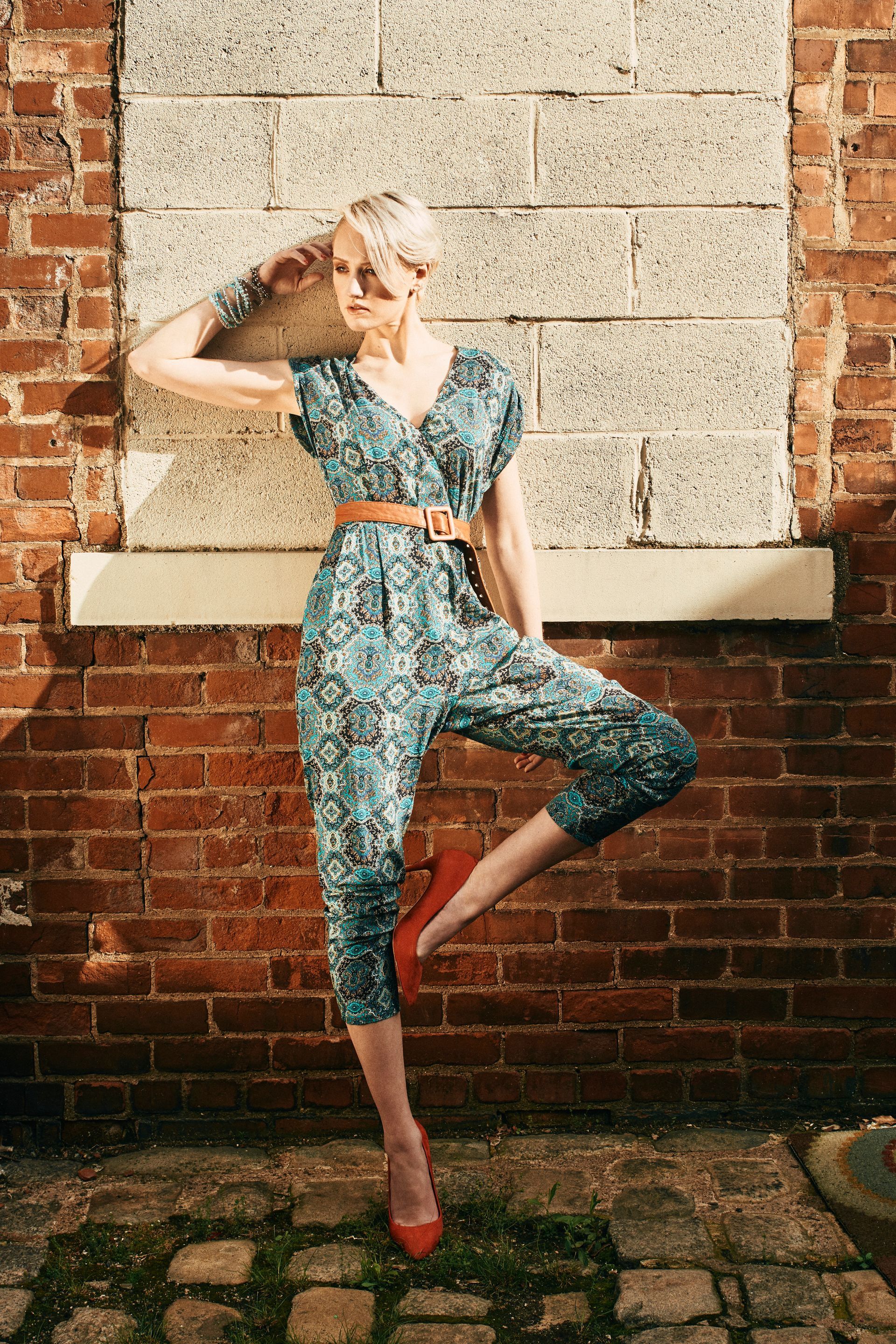 A woman in a blue jumpsuit is standing on one leg in front of a brick wall.