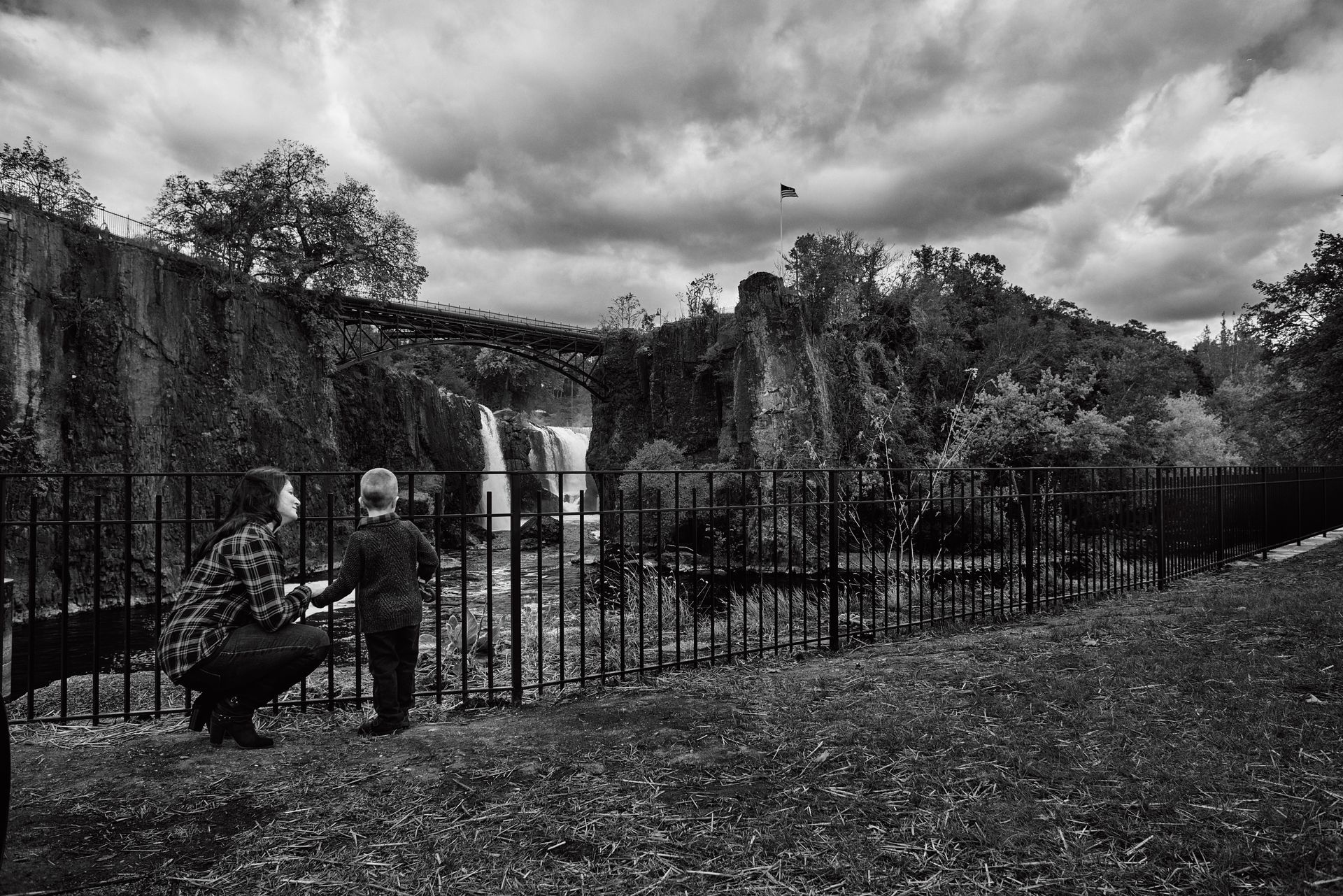 A black and white photo of two people standing next to a fence looking at a waterfall.