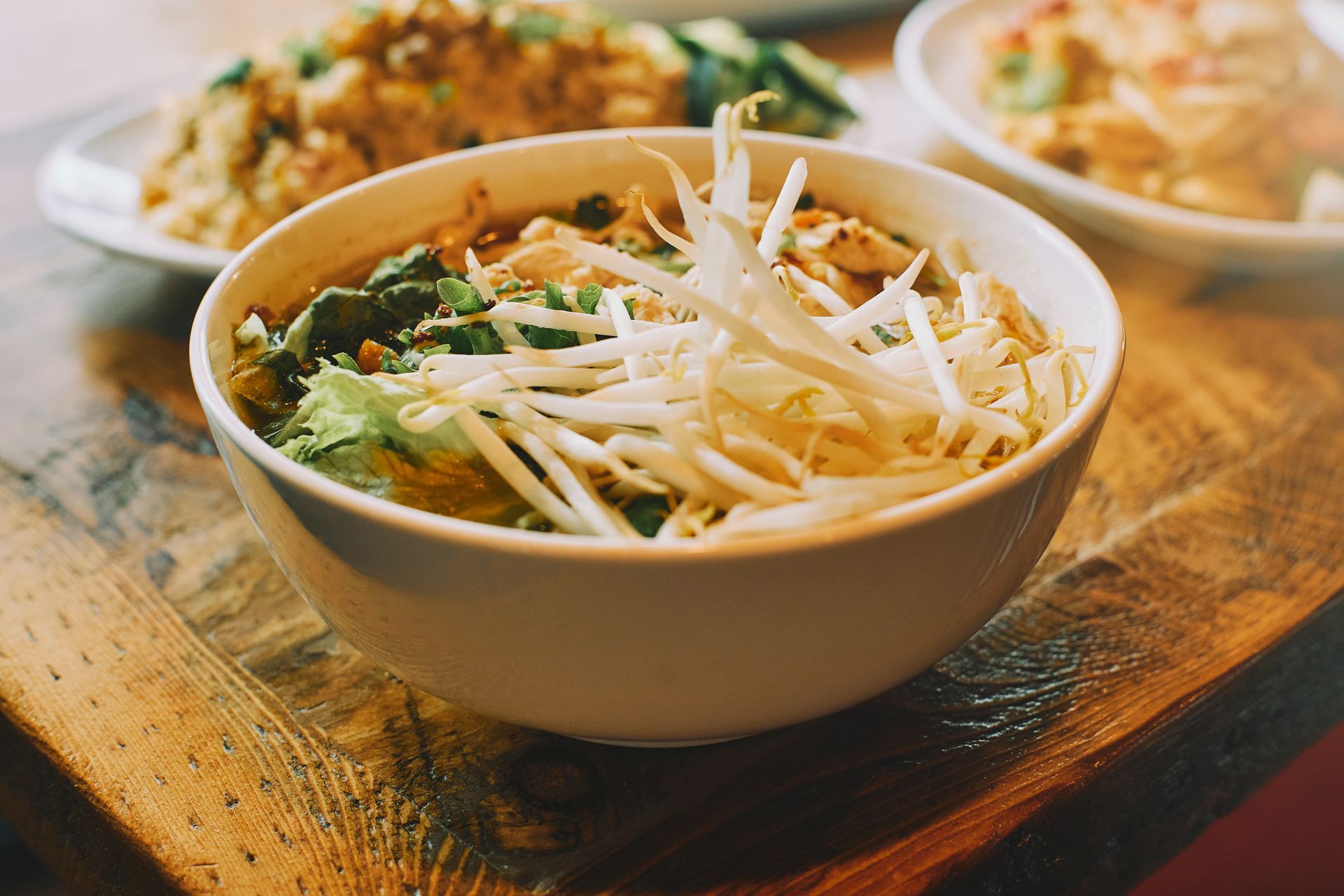 White bowl of noodle soup with bean sprouts on a wooden table. Other dishes are in the blurred background.
