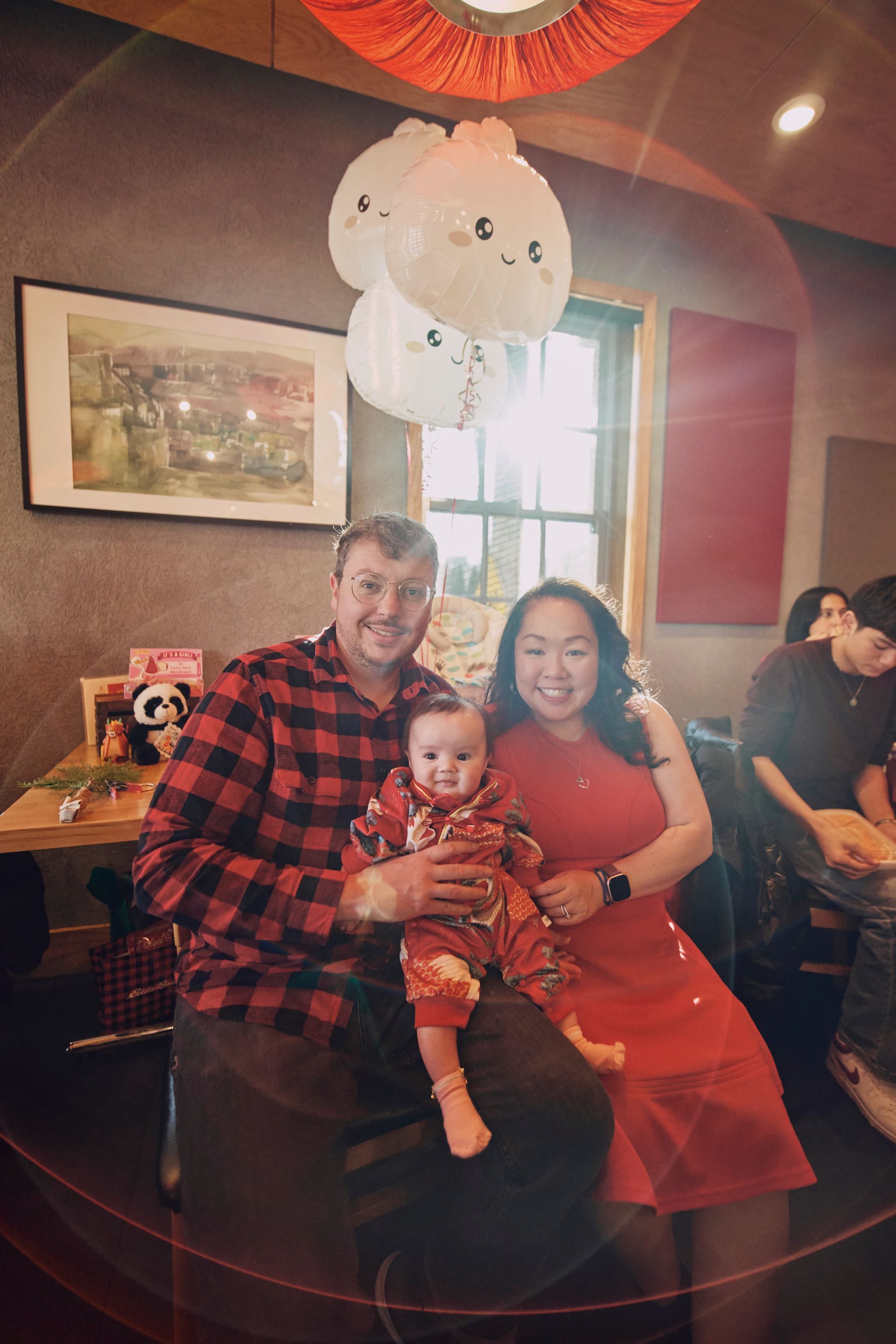 Family smiling at a camera, seated indoors. The baby is in the father's lap. The woman is in a red dress.