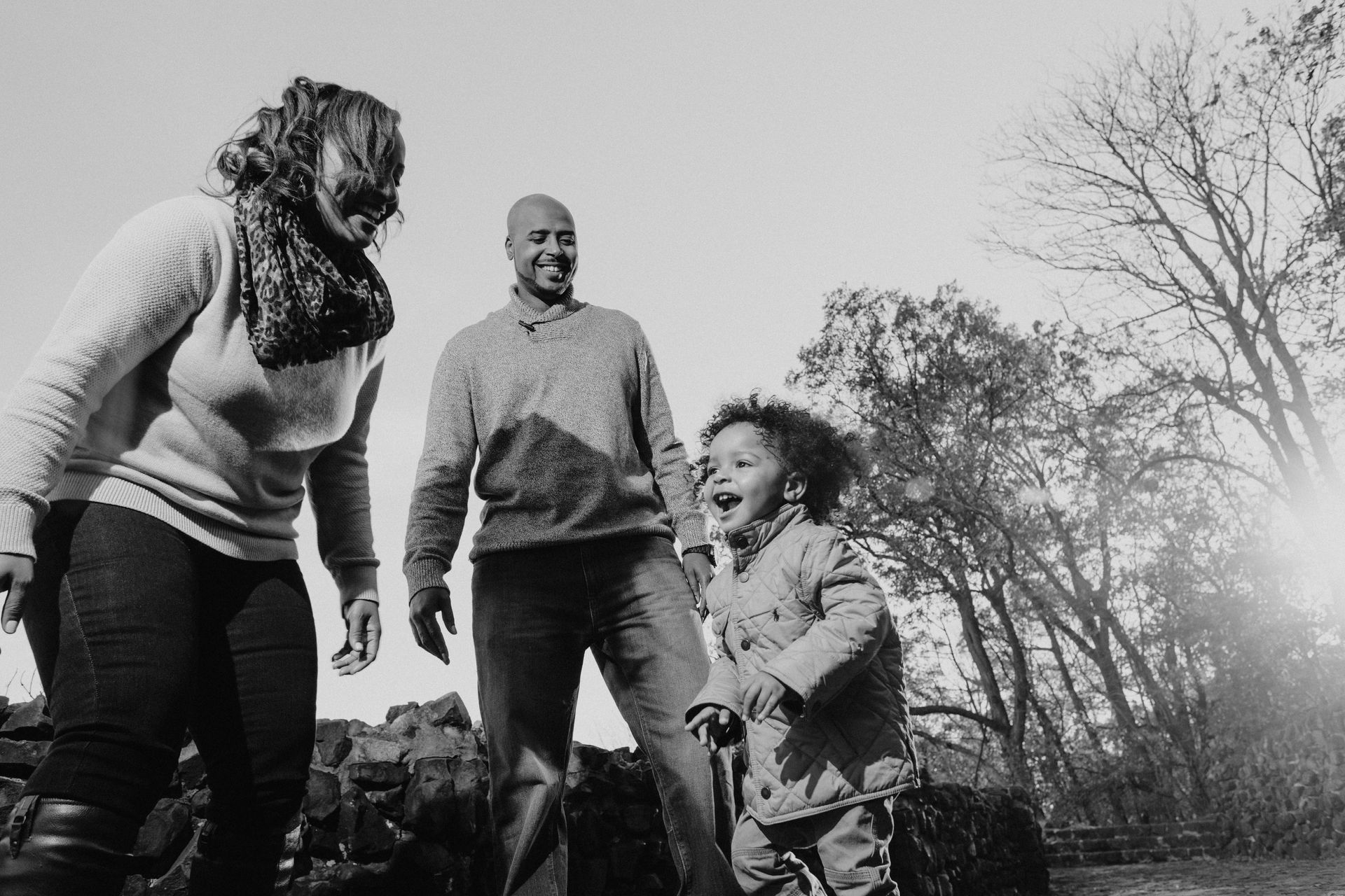 Family of three smiles outdoors in bright sunlight.