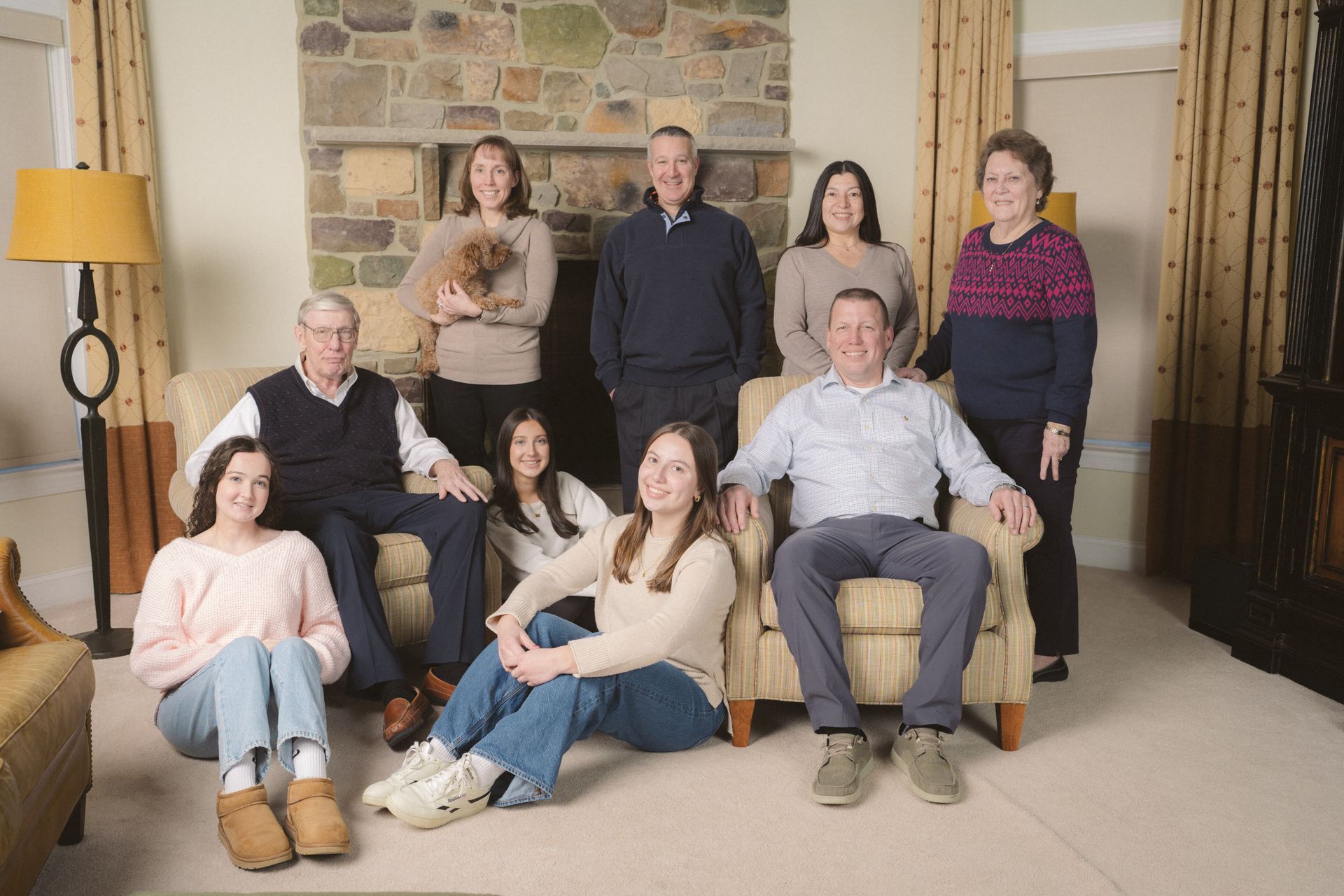 Family portrait in a living room. People sit and stand, a dog is held. Stone fireplace, neutral colors.