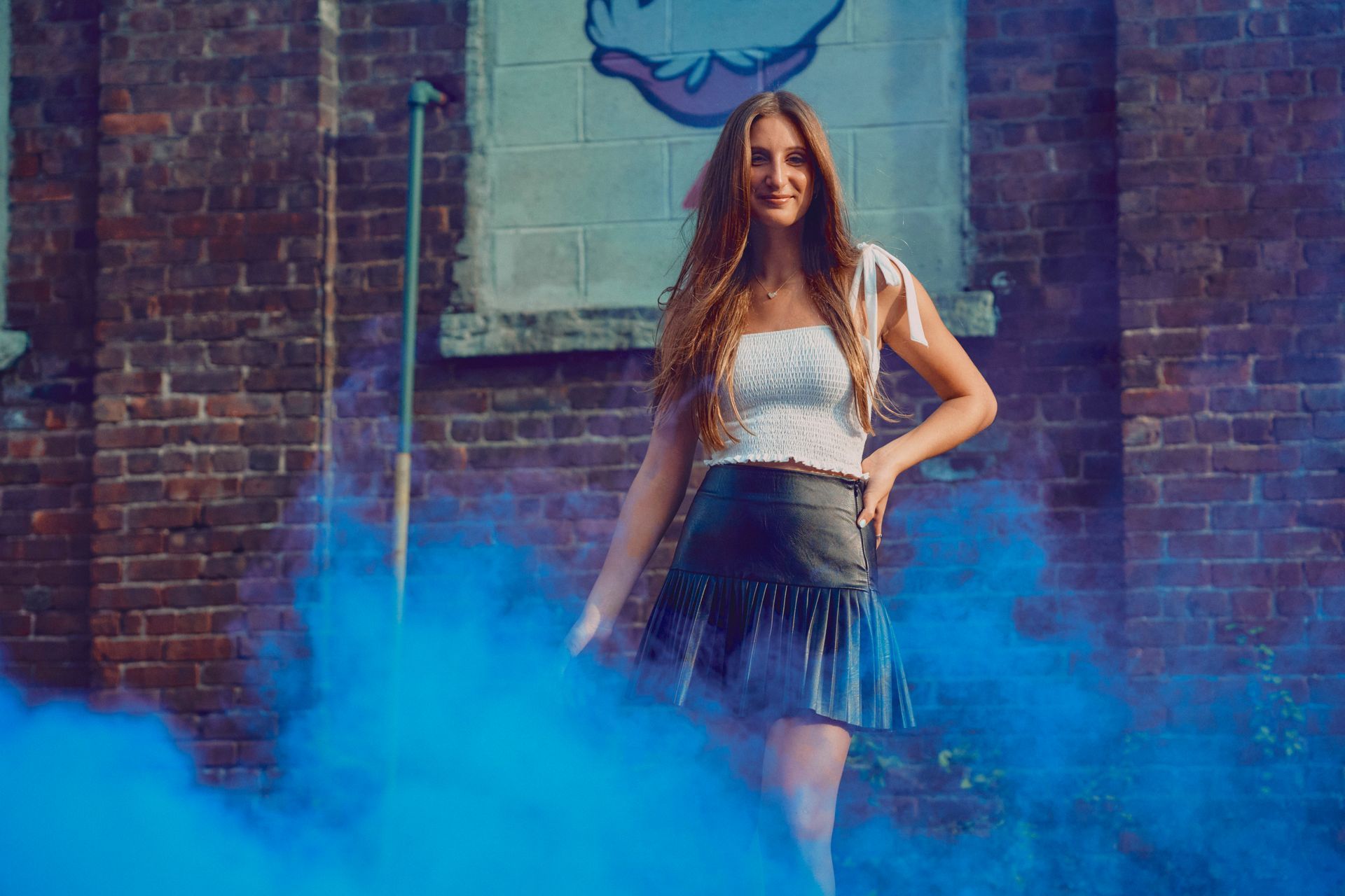 A woman is standing in front of a brick wall surrounded by blue smoke.