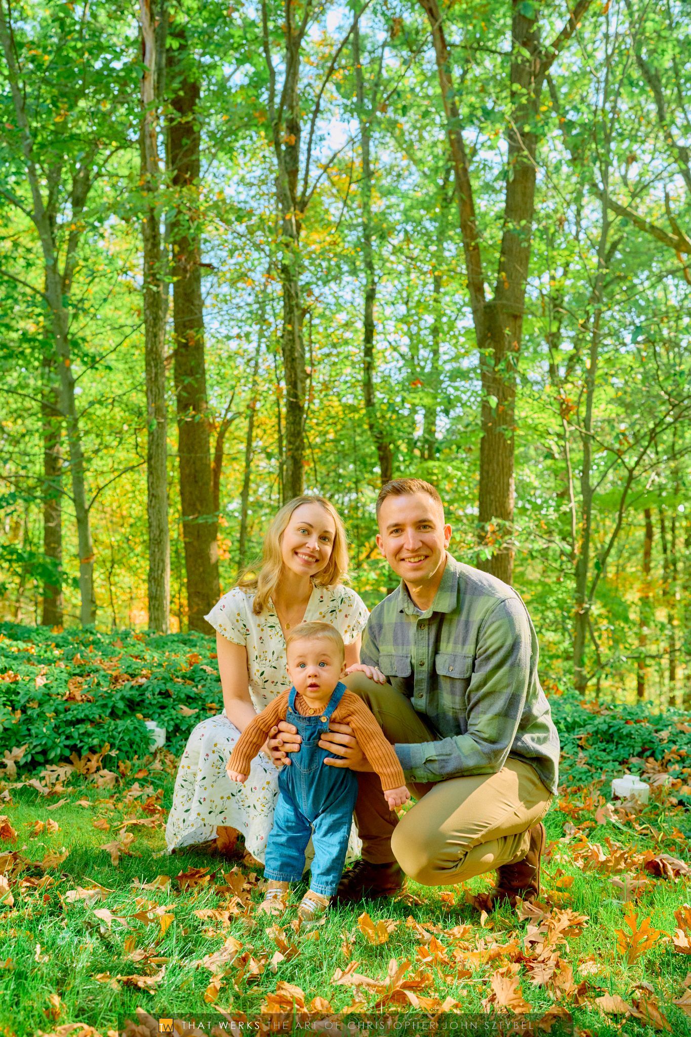 Family of three in a forest setting; parents smile, holding a toddler wearing blue overalls.