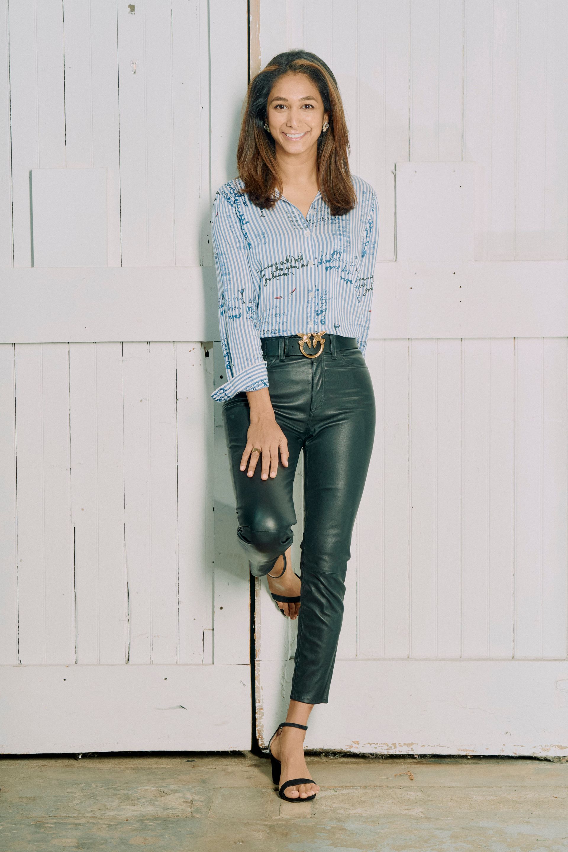 A woman in a striped shirt and leather pants is leaning against a white wall.