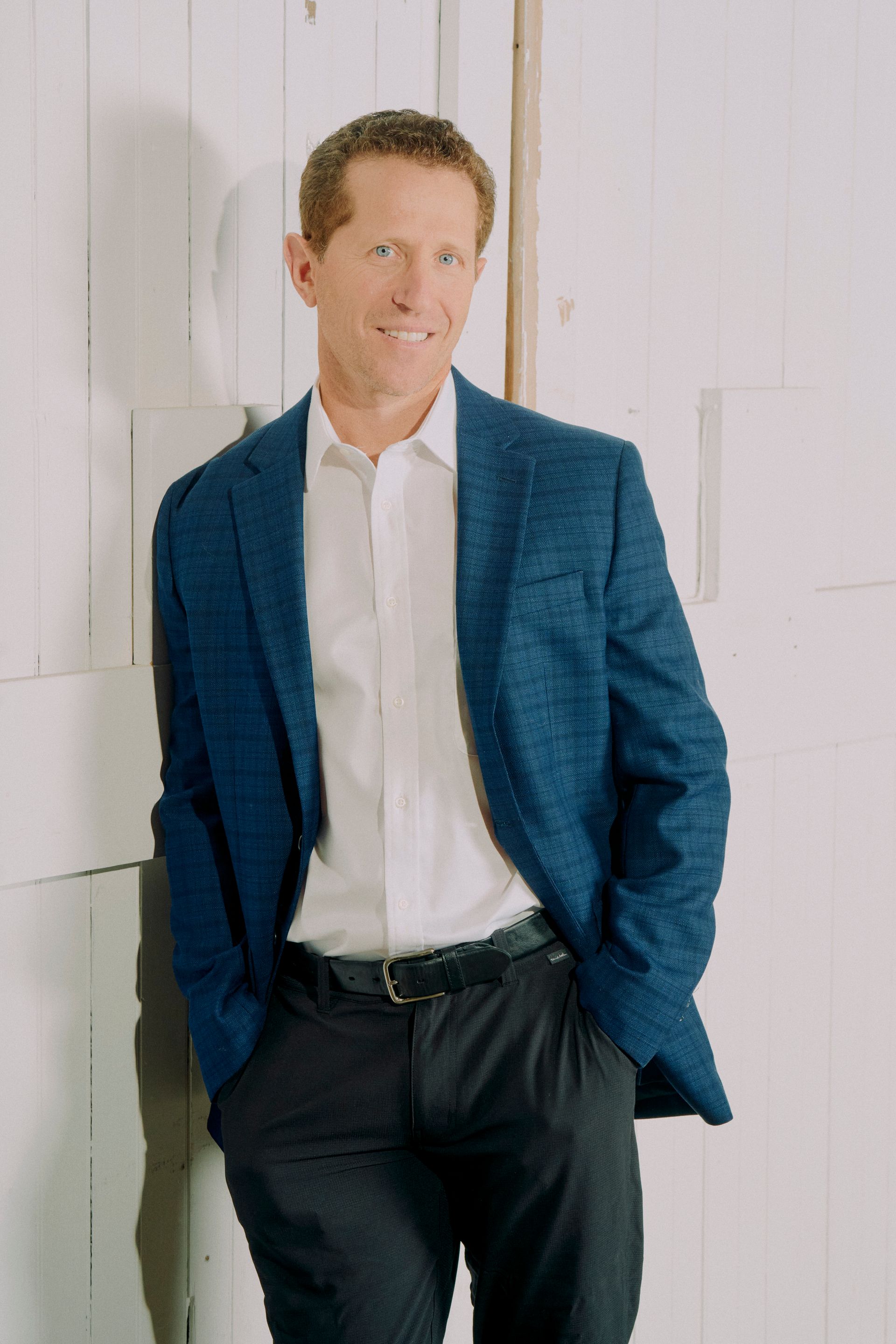 A man in a blue jacket and white shirt is leaning against a white wall.