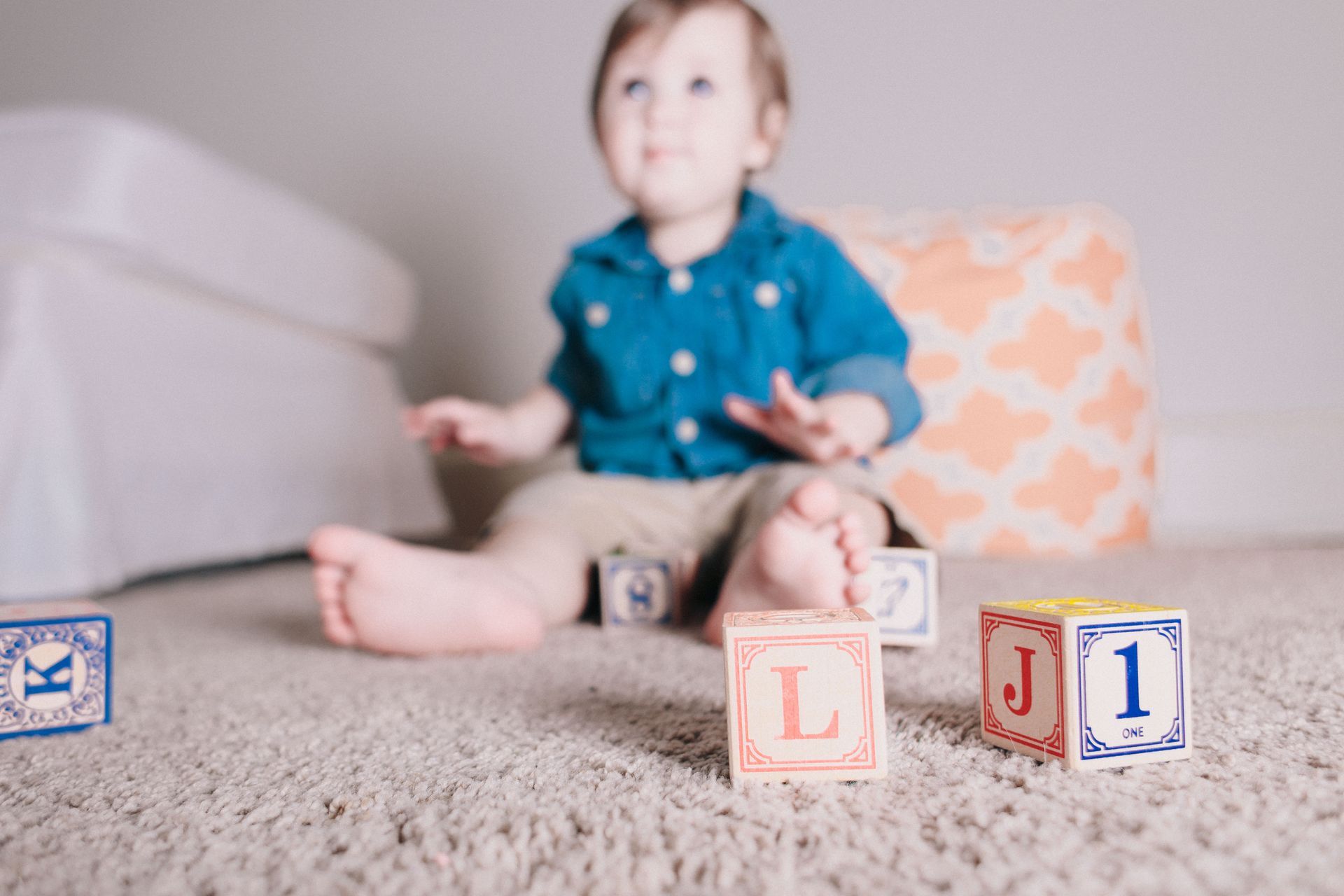 A toddler sits on a carpet playing with alphabet blocks, looking up.
