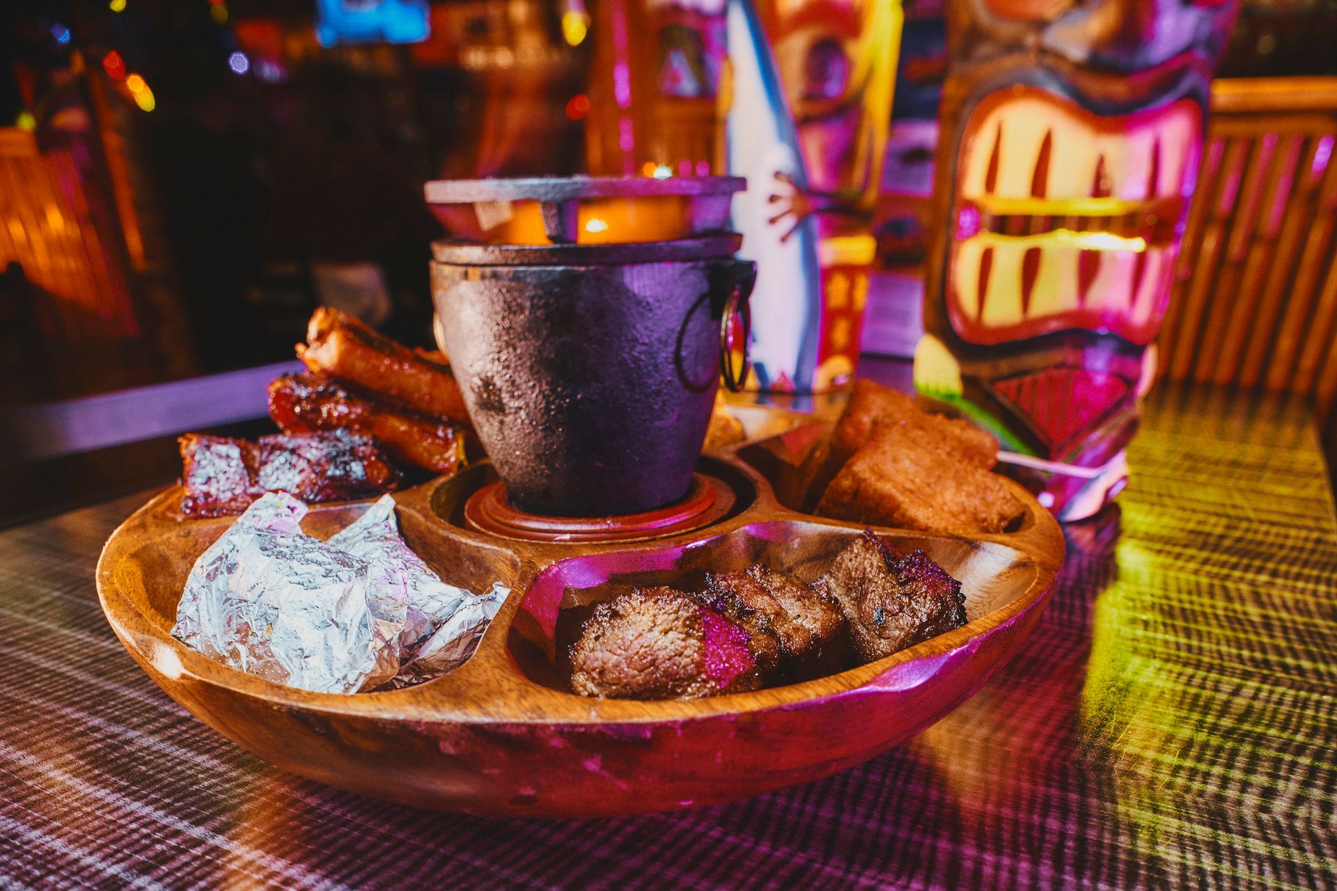Wooden serving platter with various meats, lit pot in center, tiki bar setting.