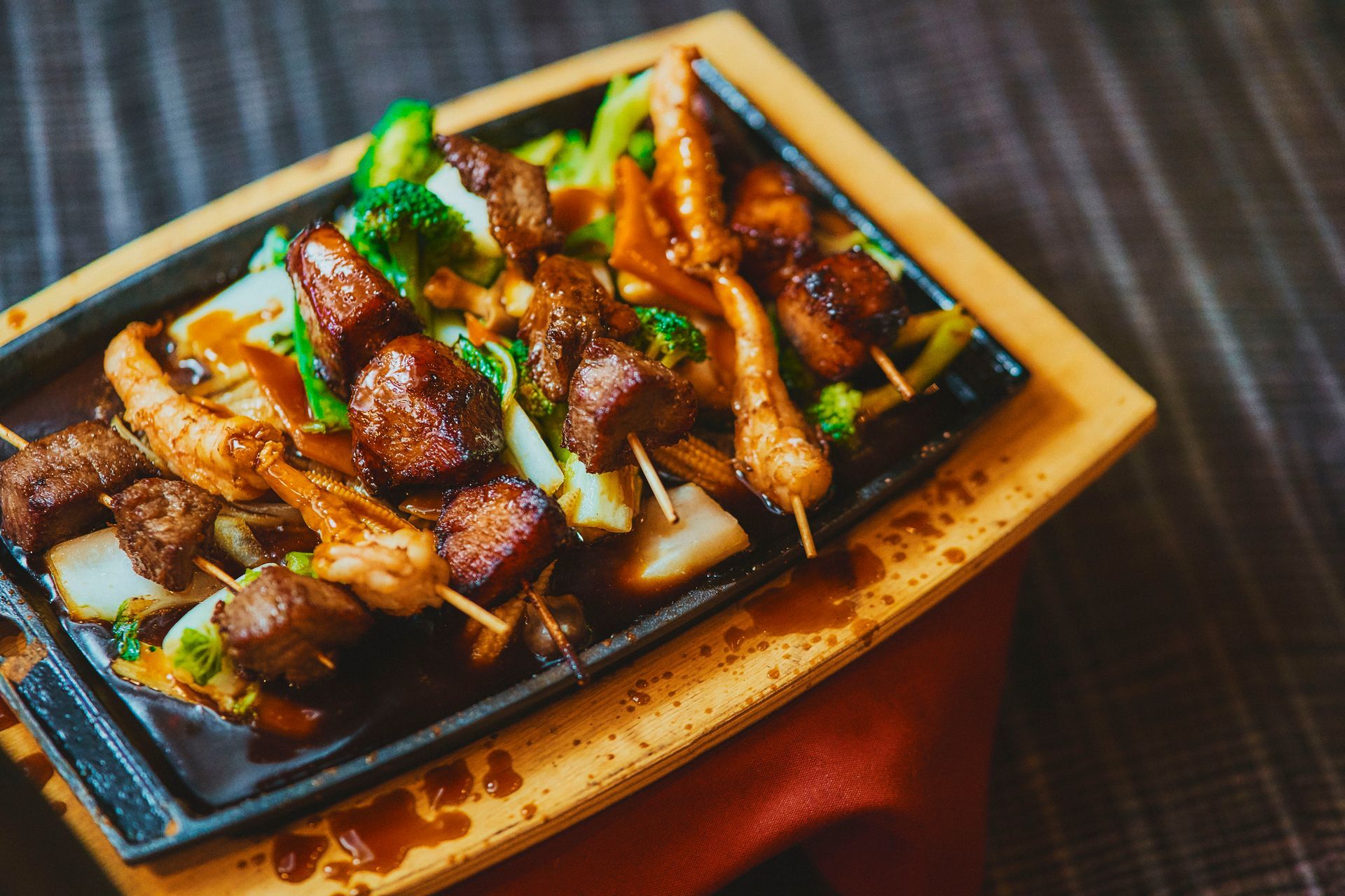 Skewered meat and vegetables on a sizzling platter, drizzled with sauce; served on a wooden tray.