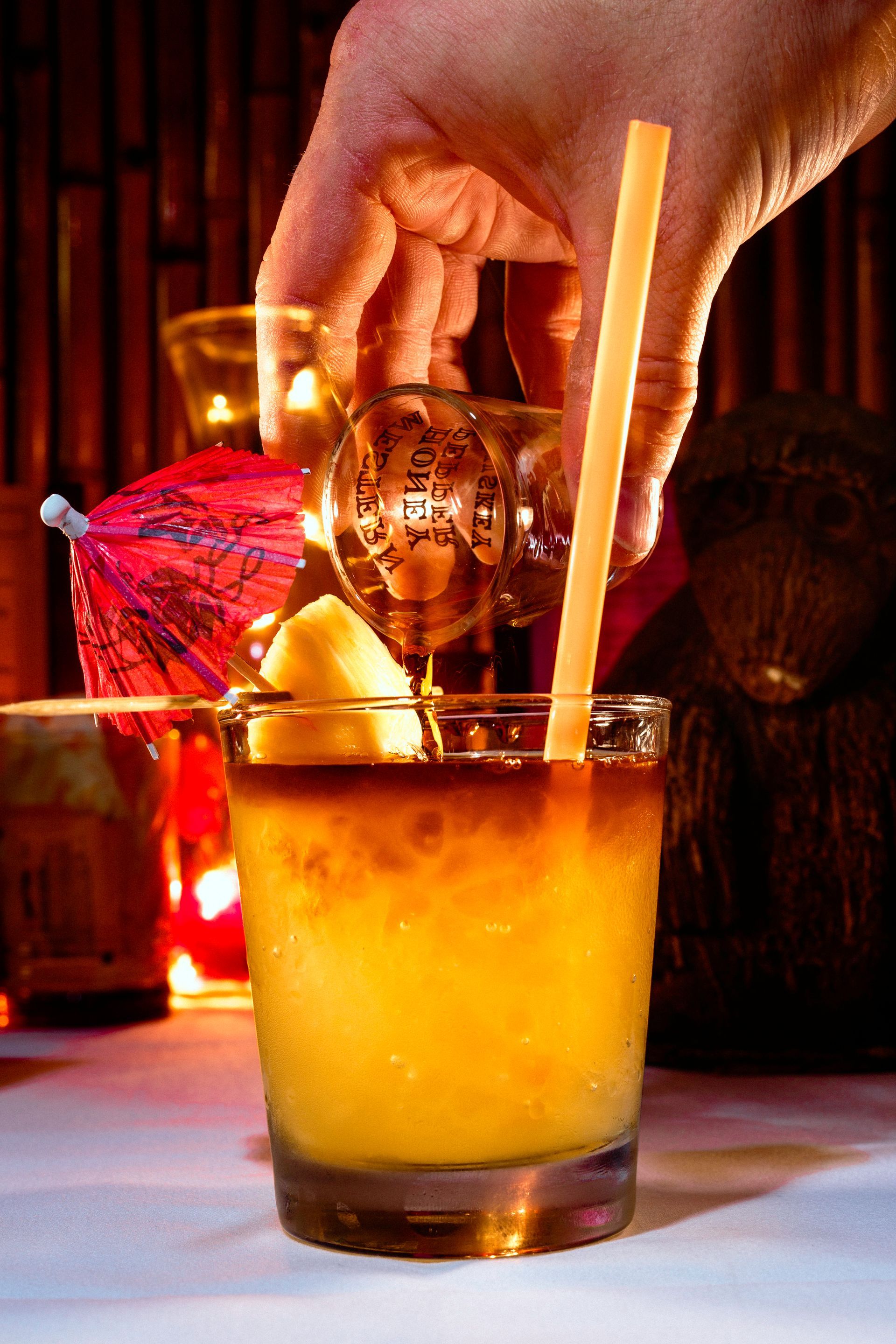 Hand pouring liquid into cocktail glass with ice, bamboo straw, garnished with fruit.
