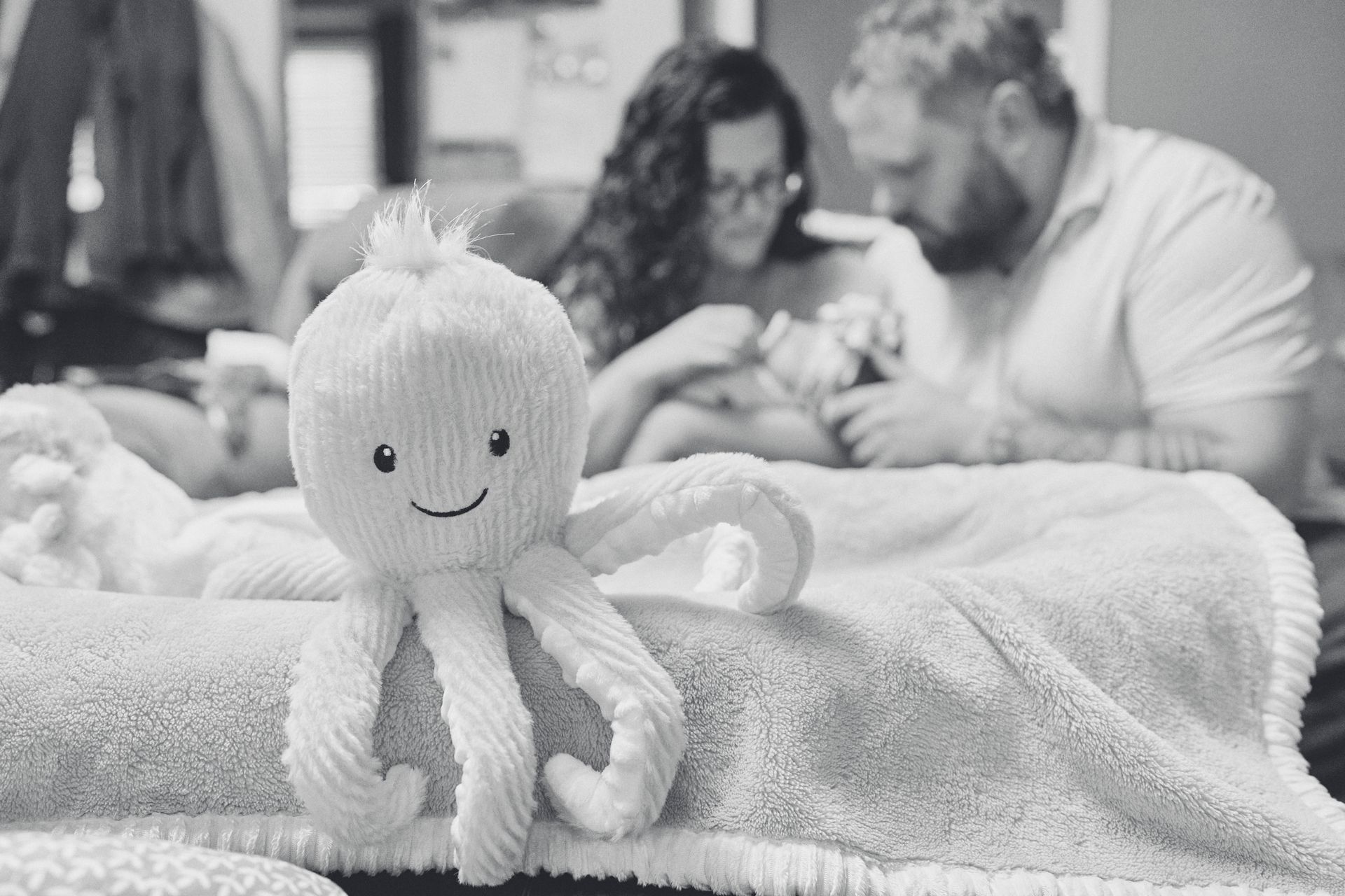 Stuffed octopus toy on a blanket, with parents looking at a newborn.