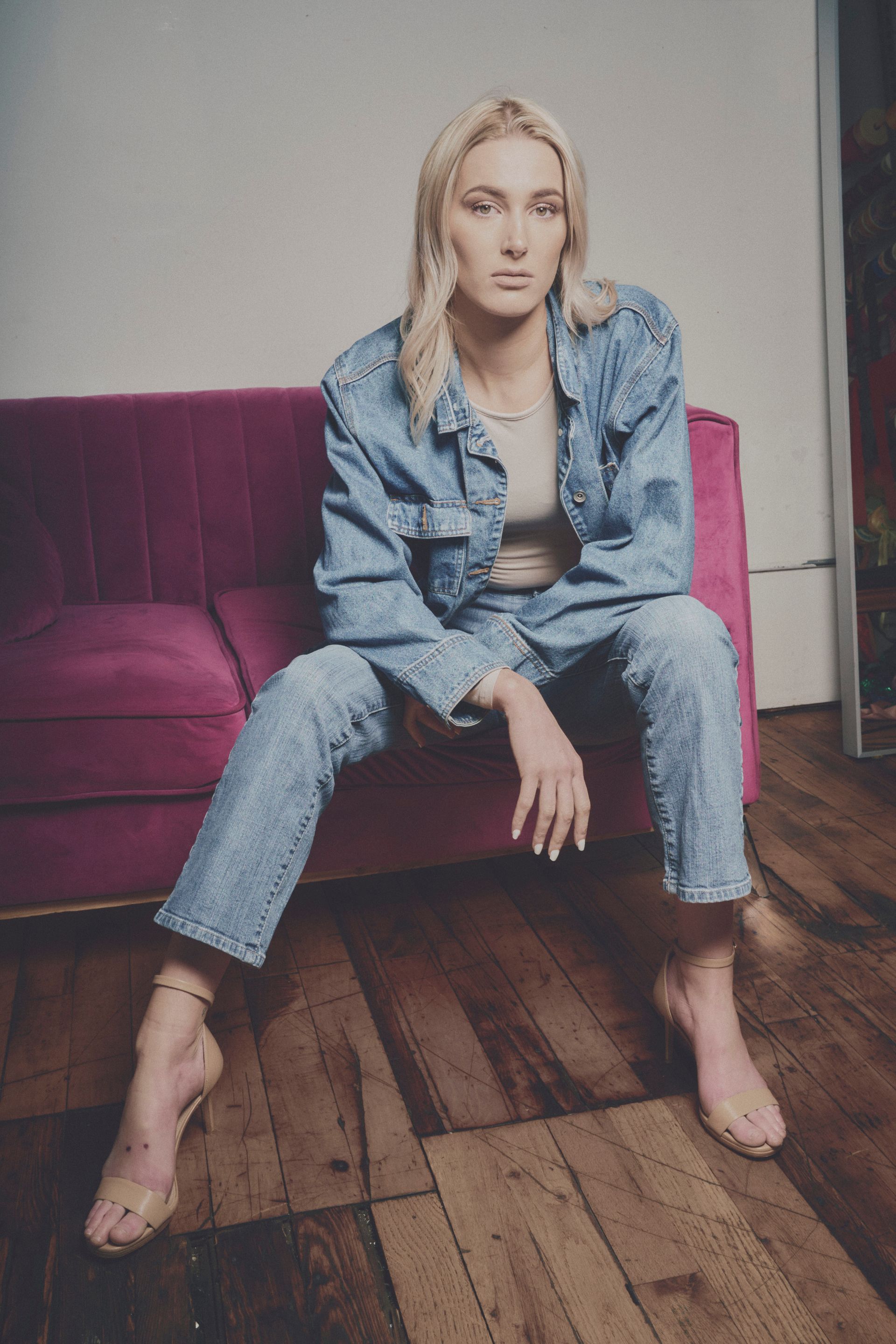 A woman in a denim jacket and jeans is sitting on a pink couch.