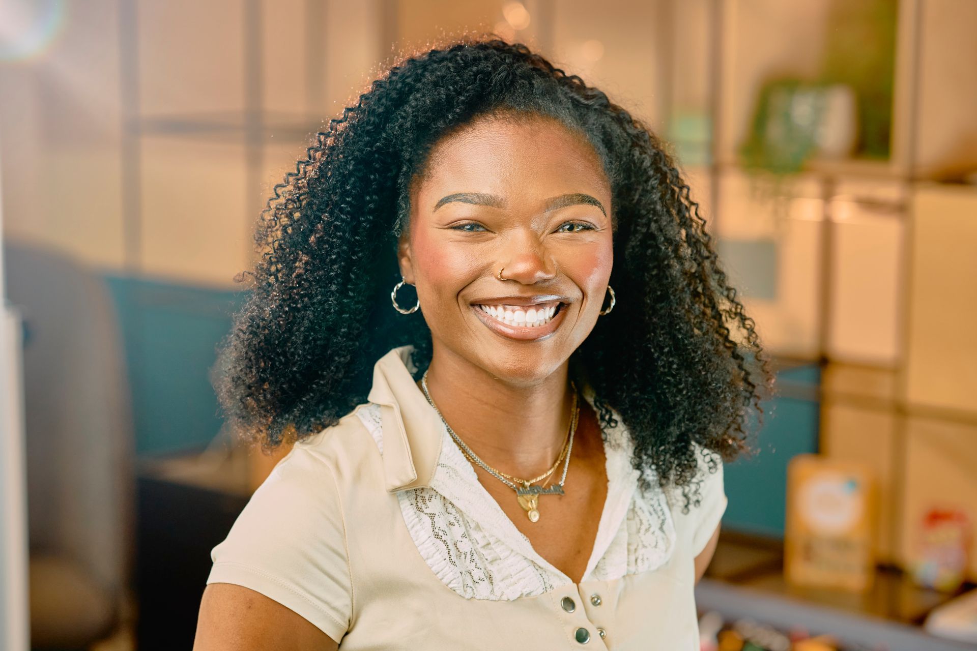 Smiling woman with curly hair in a bright office, wearing a white blouse and necklace