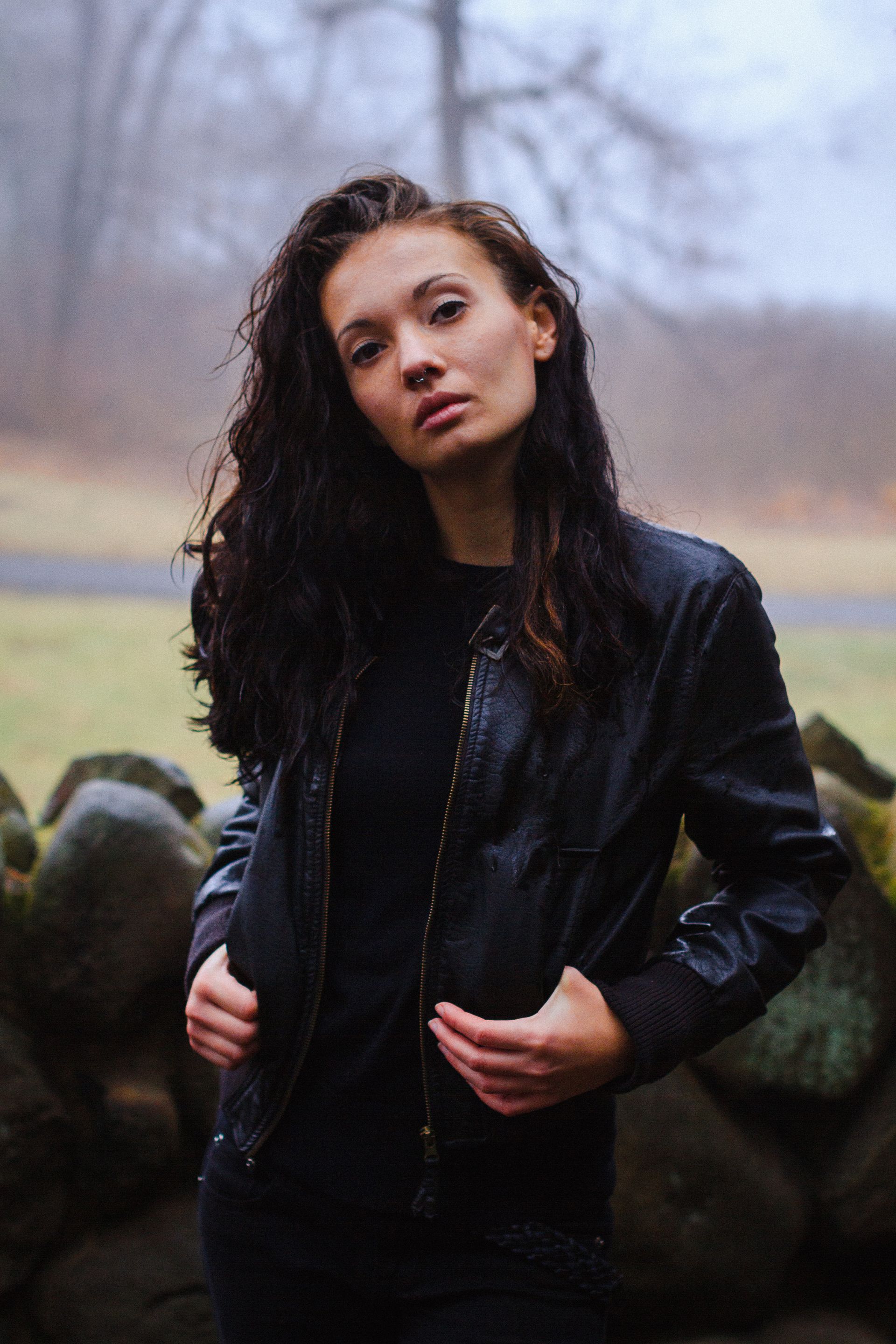 A woman in a black leather jacket is standing in front of a stone wall.