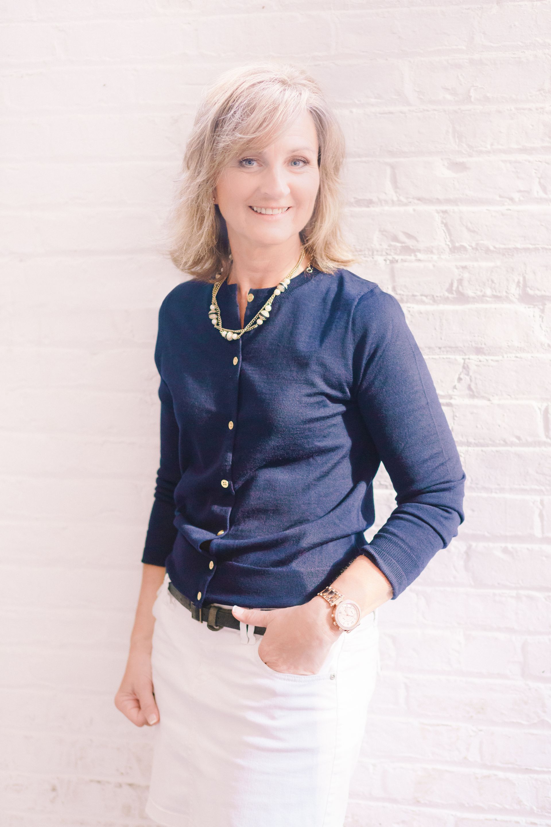 A woman wearing a blue cardigan and white skirt is standing in front of a white brick wall.