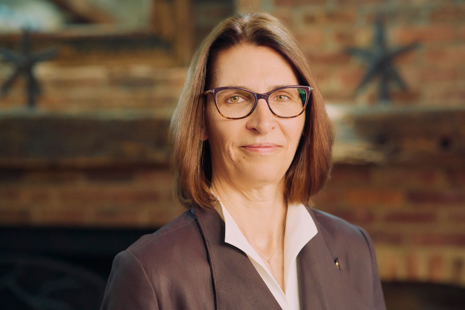 Portrait of a woman with glasses in a dark blazer, standing before a blurred wooden wall