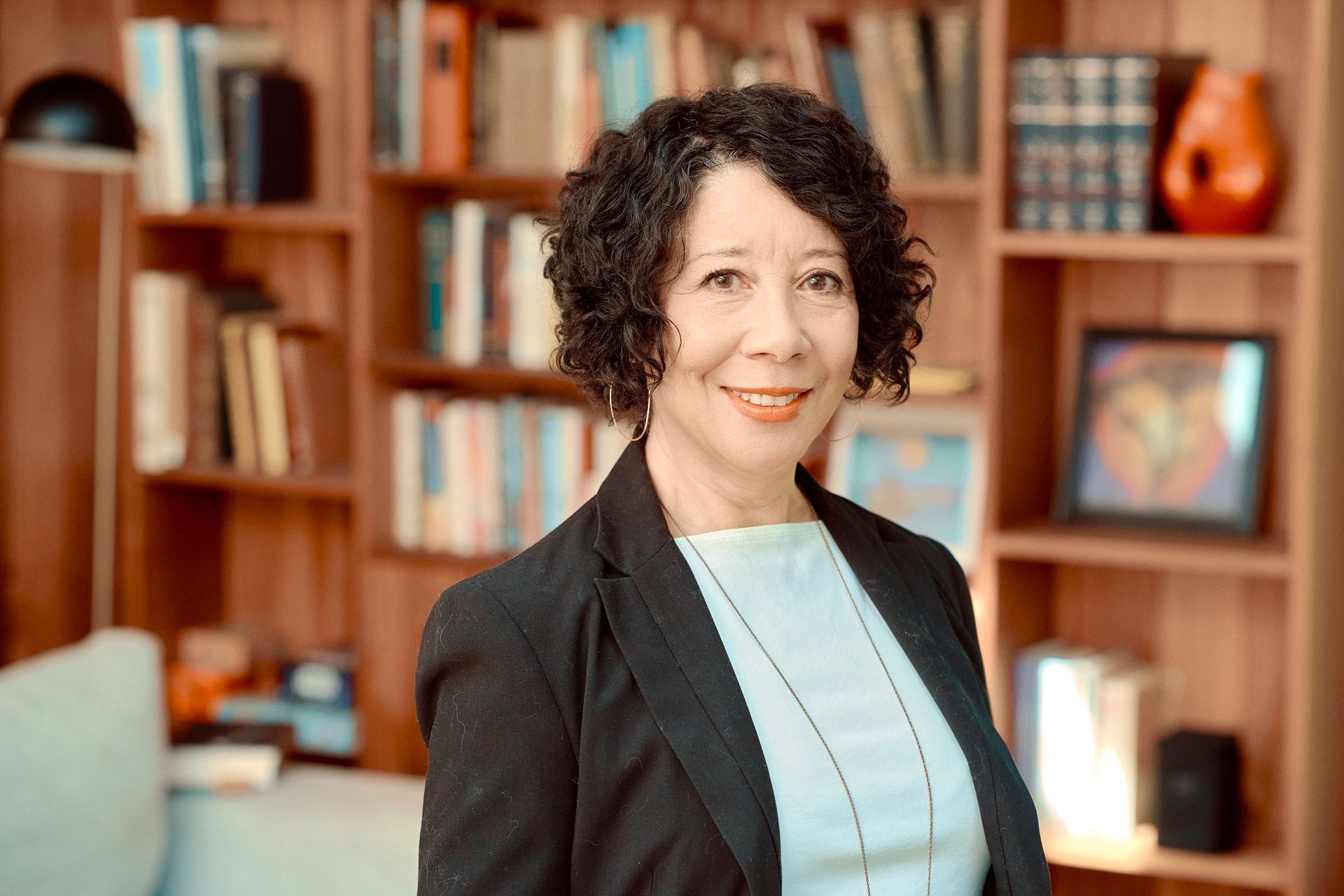 Woman in a blazer smiles in front of a bookcase, a lamp, and artwork.