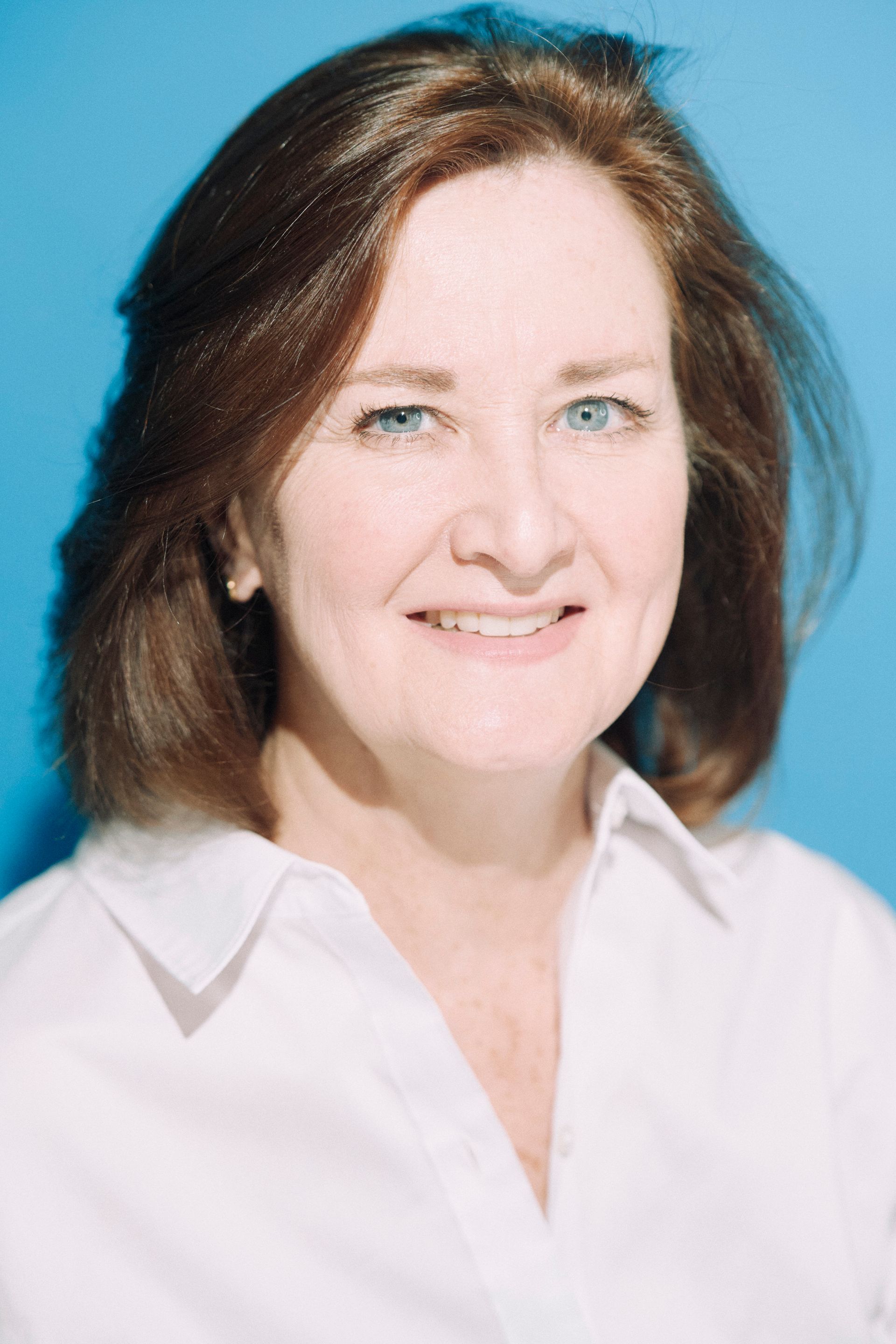 A woman in a white shirt is smiling in front of a blue background.