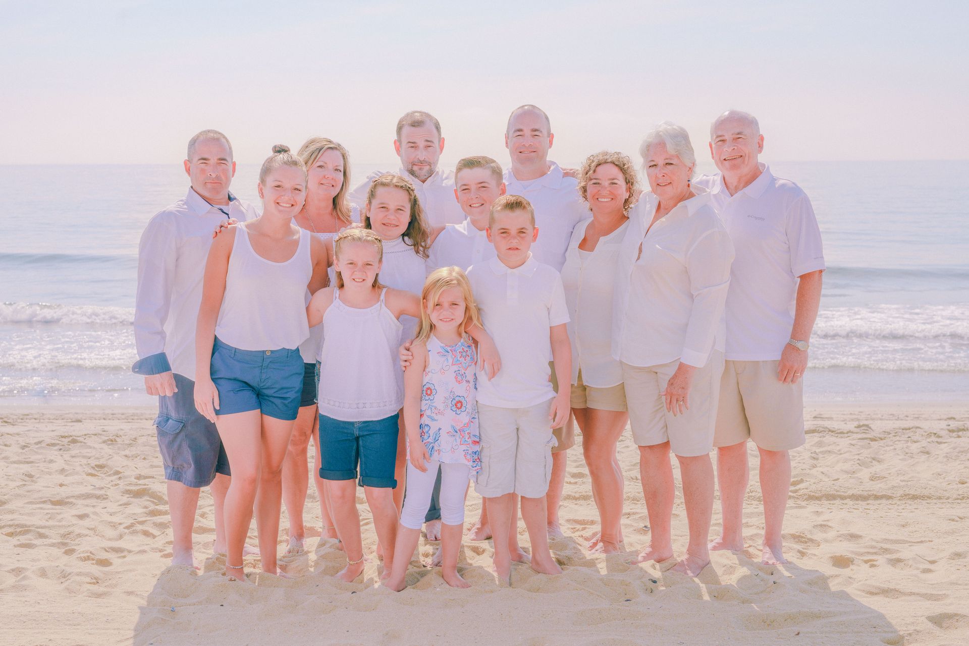 Large family group in white outfits standing on a sandy beach, smiling. The ocean is in the background.