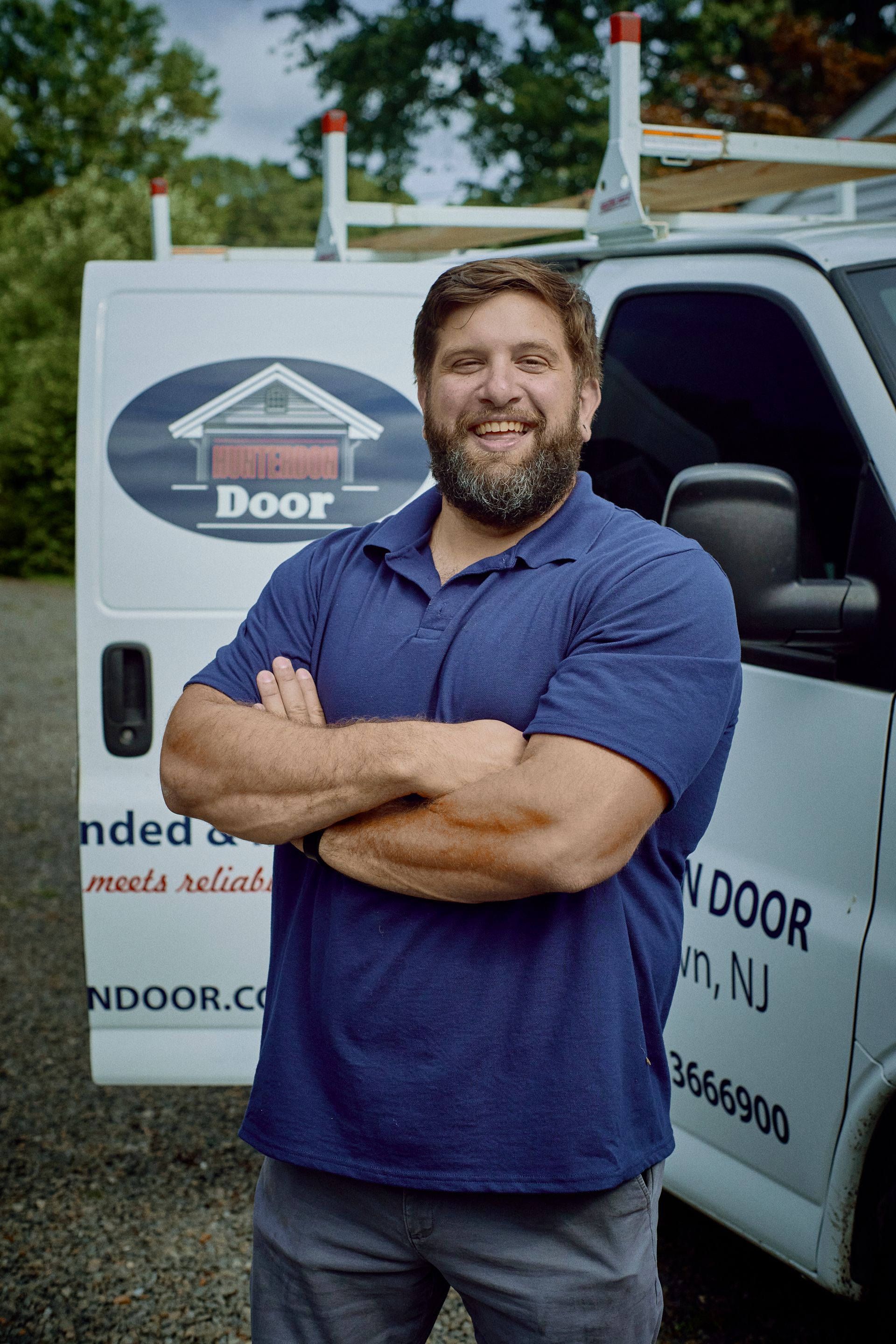 Man with crossed arms in front of white van with company logo. He's wearing blue and smiling outdoors.