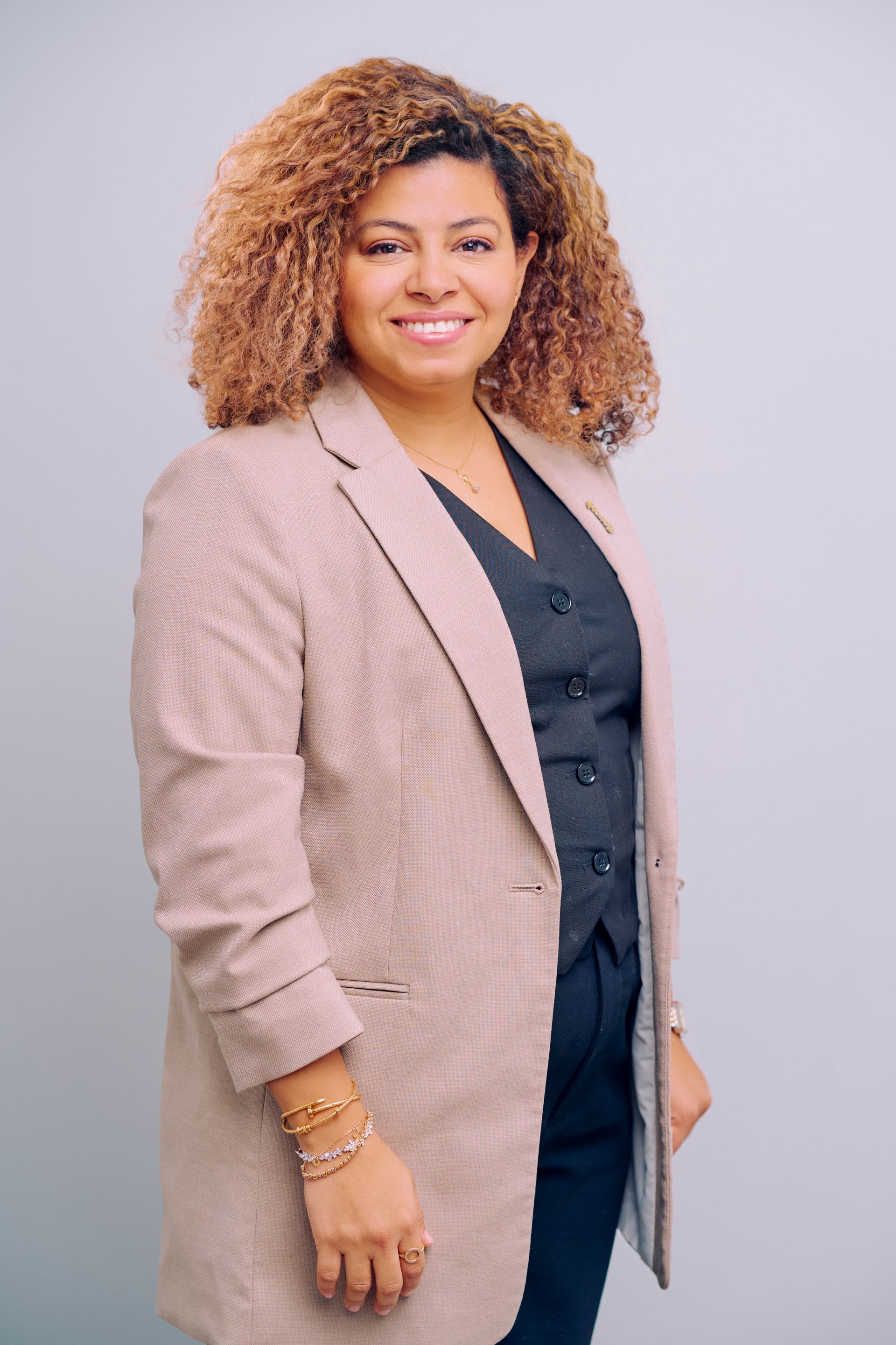 Woman in blazer and vest smiles, standing against a gray background.