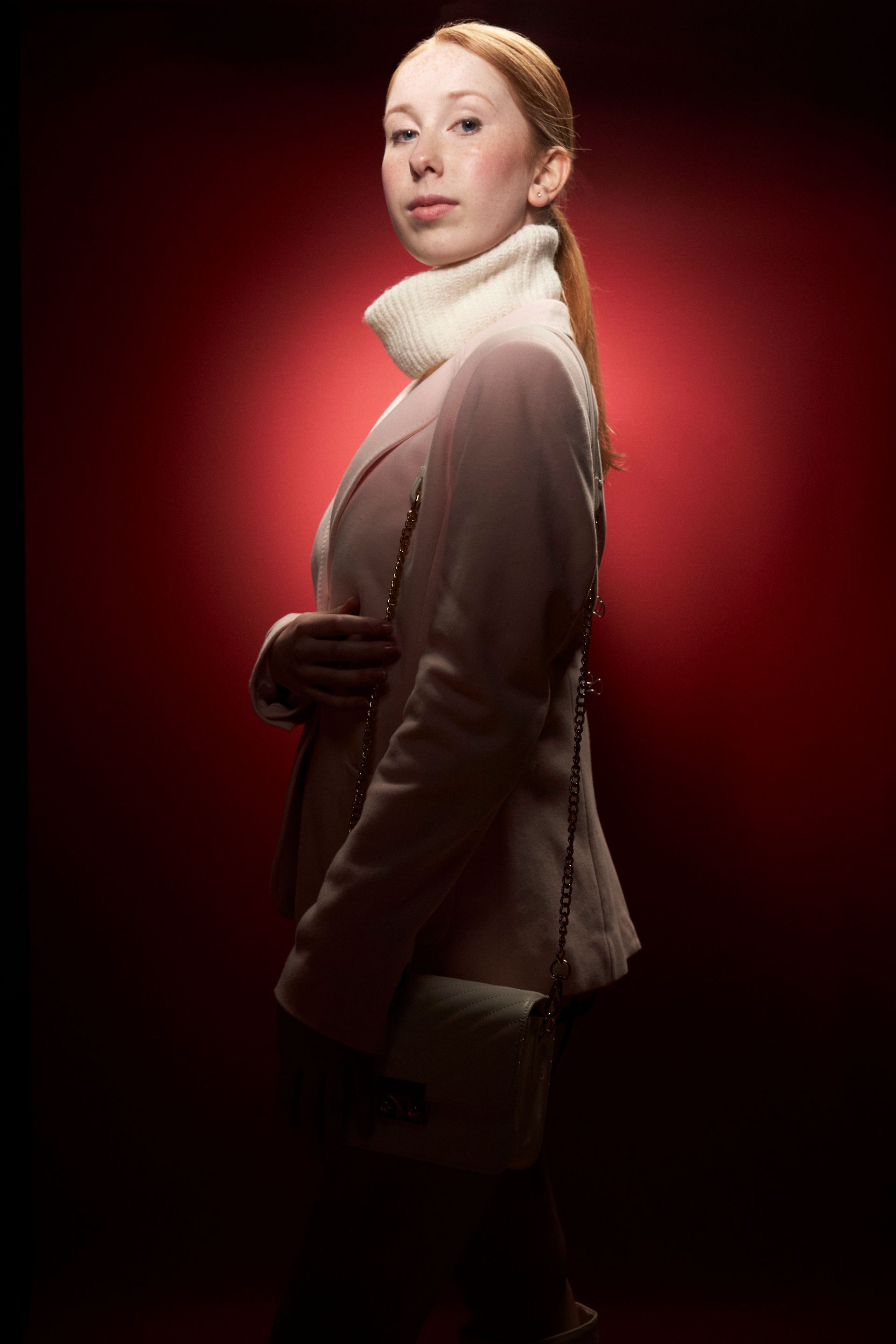 A woman in a pink coat and white turtleneck is standing in front of a red background.