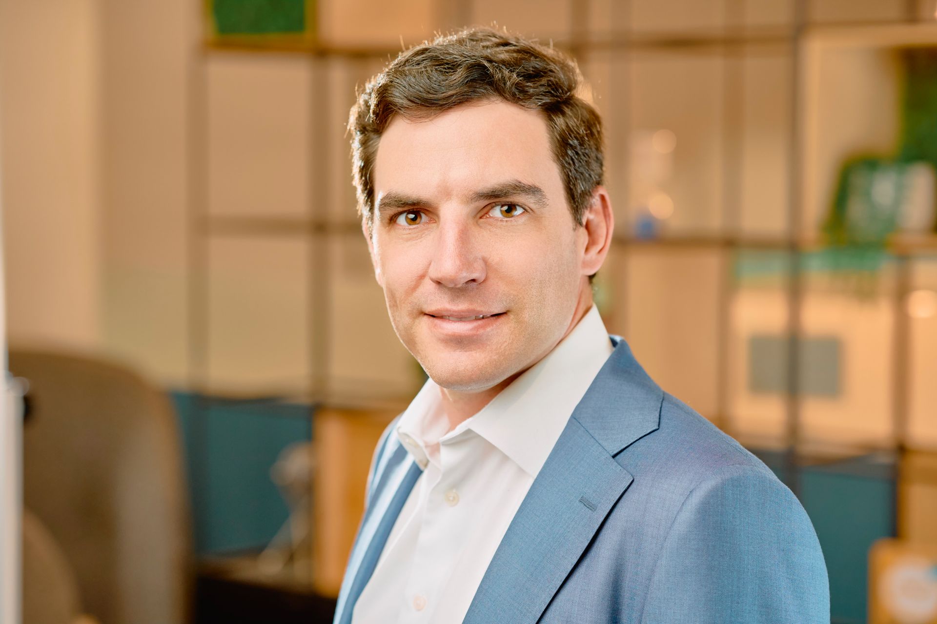 Portrait of a man in a light blue suit smiling in a modern office setting