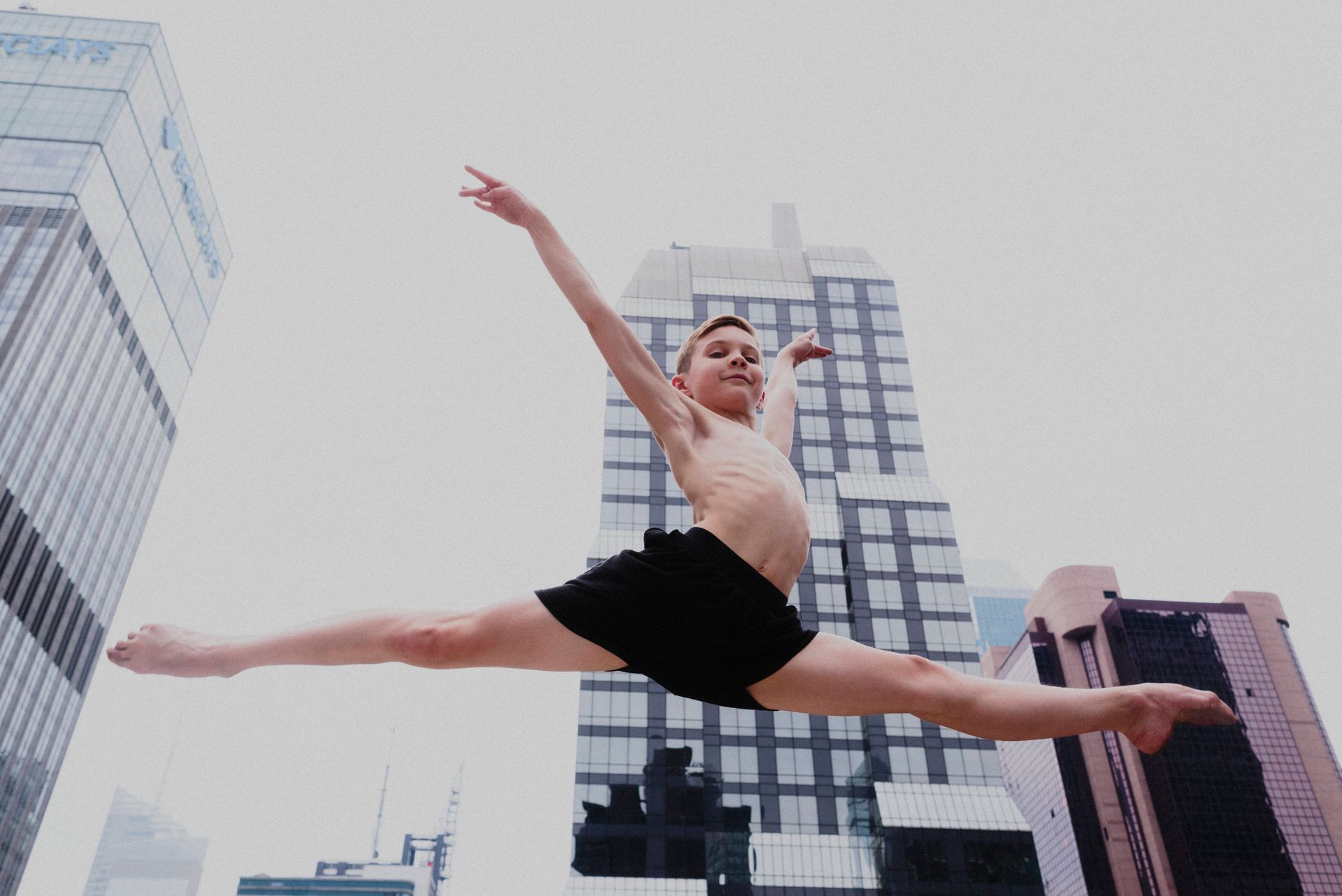 A young boy is doing a split in the air in front of tall buildings