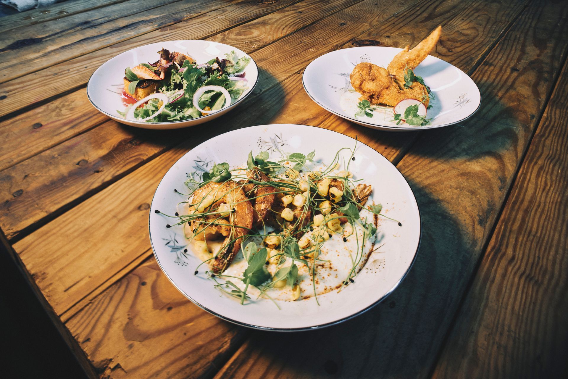 Three plates of food on a wooden table. The dishes include salad, fried food, and garnished ingredients.