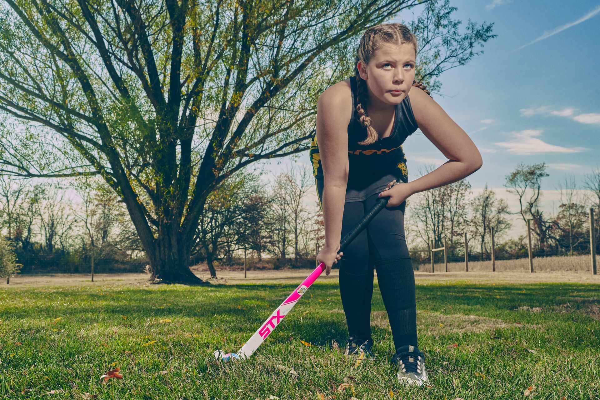 Girl in athletic wear with field hockey stick in a grassy area, preparing to play.