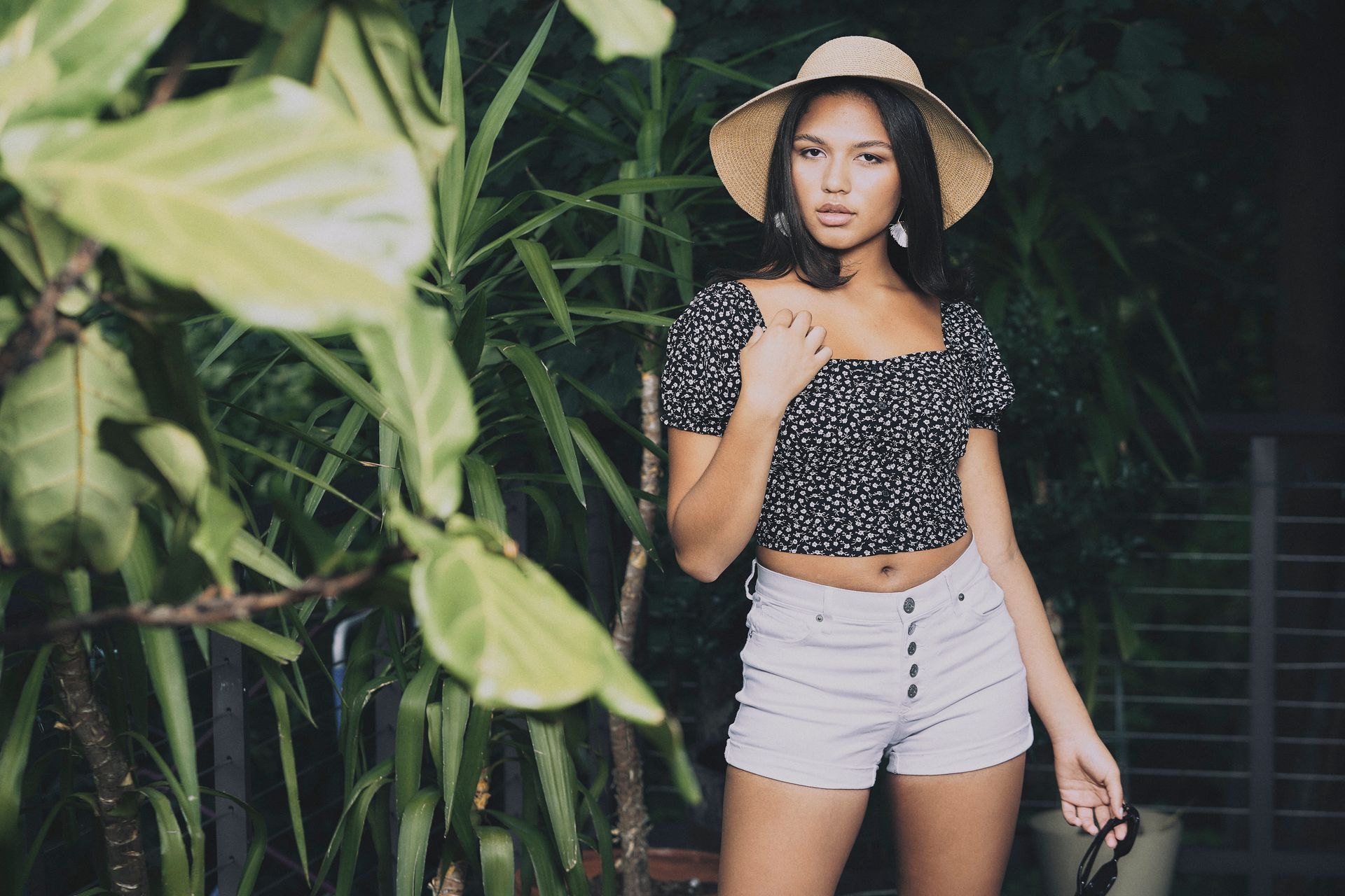 A woman wearing a hat and shorts is standing in front of a plant.