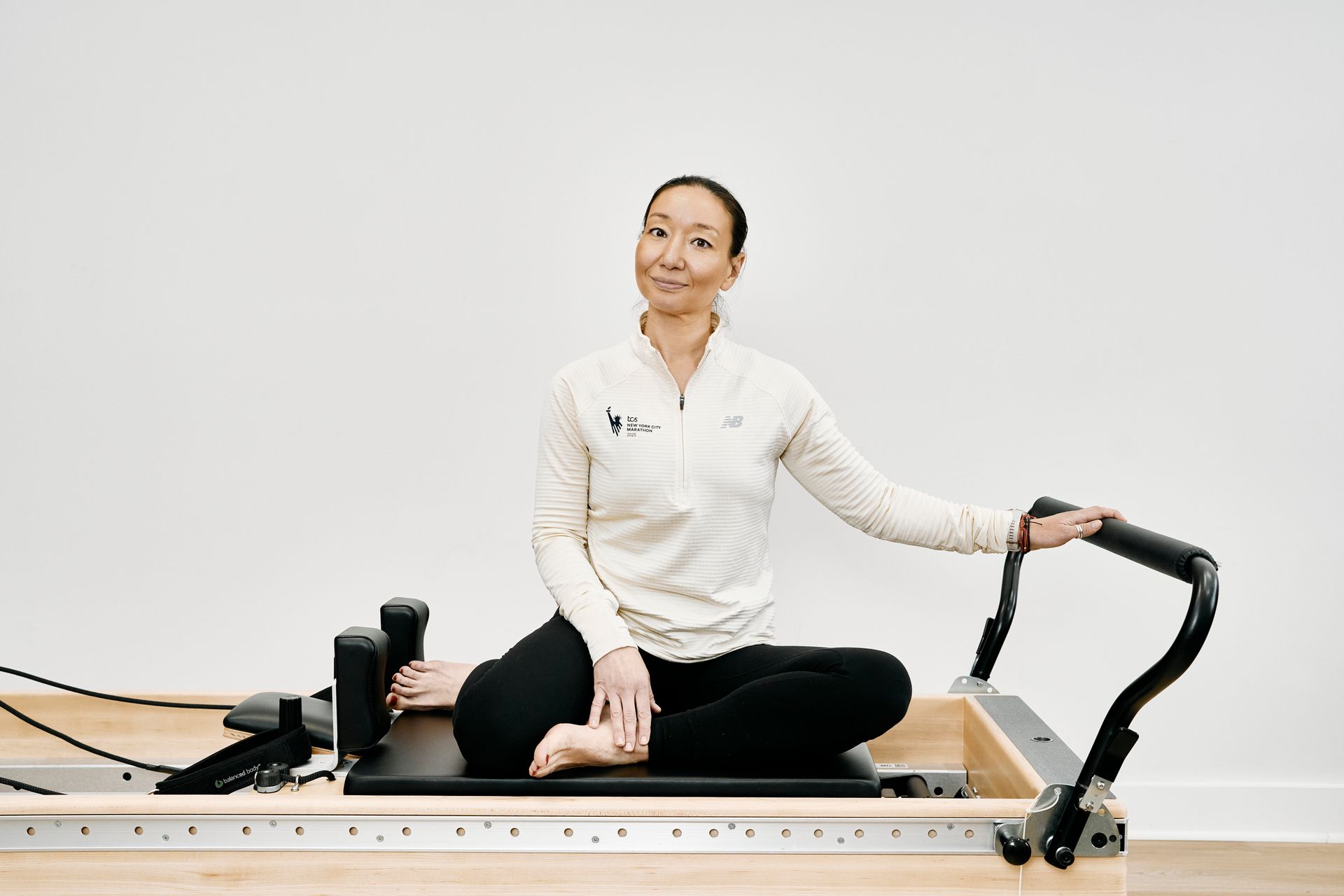 Person seated cross-legged on a Pilates reformer, holding the handlebar and smiling in a studio