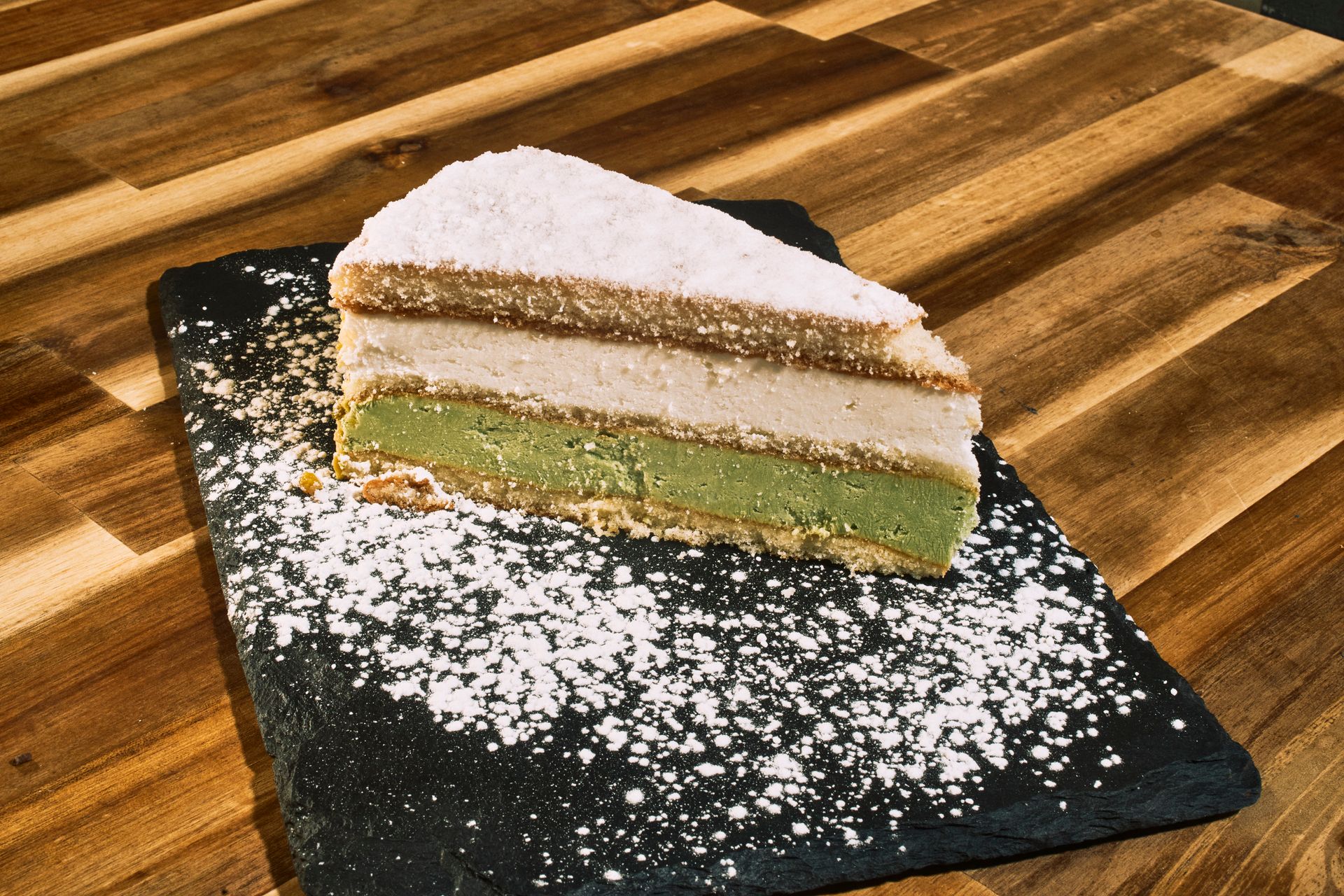 Slice of layered cake on a black slate, dusted with powdered sugar, set on a wooden surface.