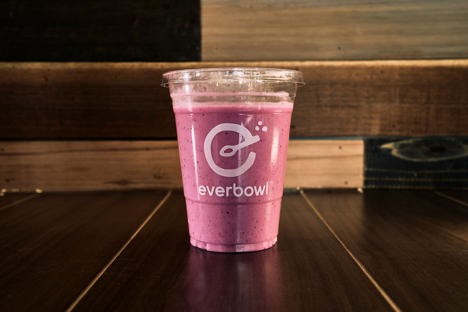 Purple smoothie in clear plastic cup on wood surface. The cup has a logo that reads 