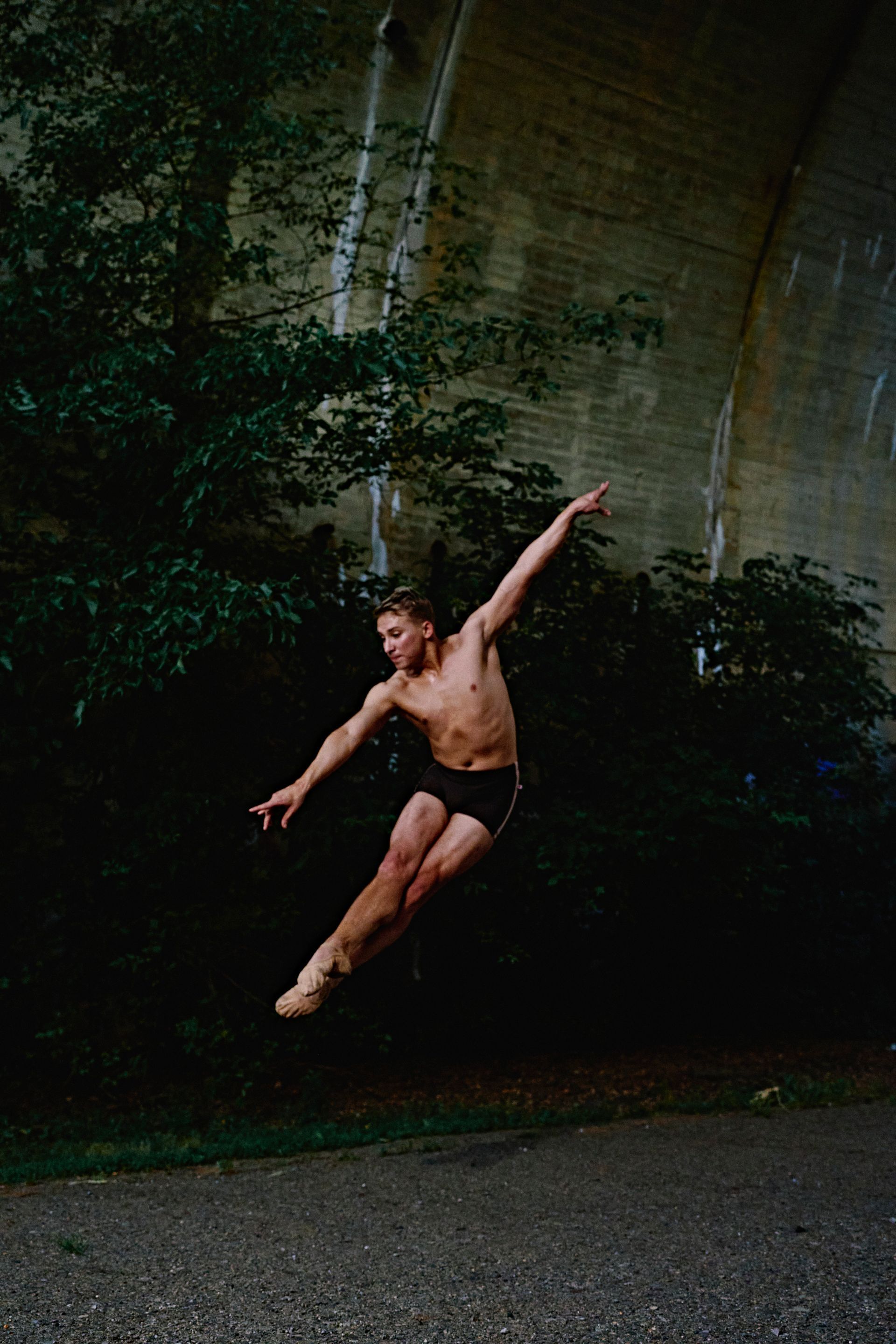 Man leaping in the air, arms extended, under a bridge. Wearing black shorts, outdoors.